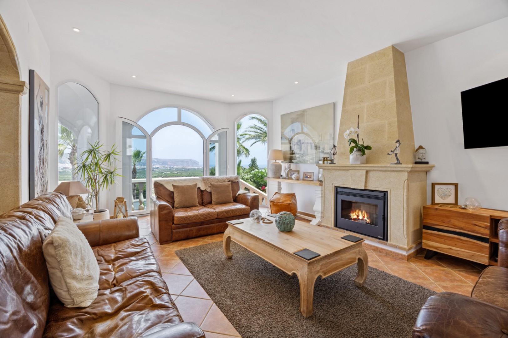Séjour élégant avec cheminée en pierre, mobilier en cuir et portes voûtées s'ouvrant sur une terrasse avec vue panoramique sur la mer.