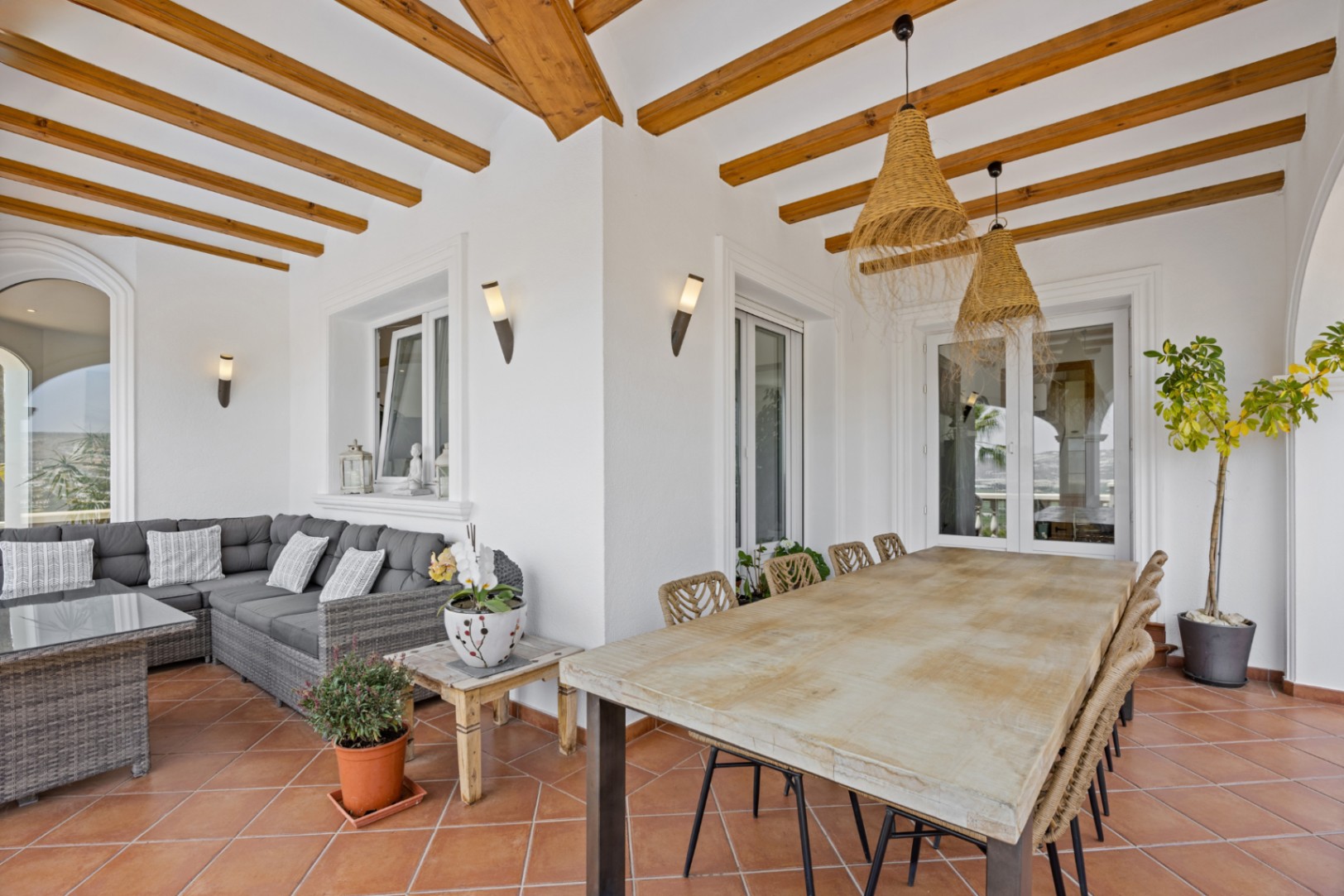 Terrasse couverte avec poutres en bois, sol en terre cuite, grande table à manger et salon de jardin en résine tressée grise sous des suspensions en paille.