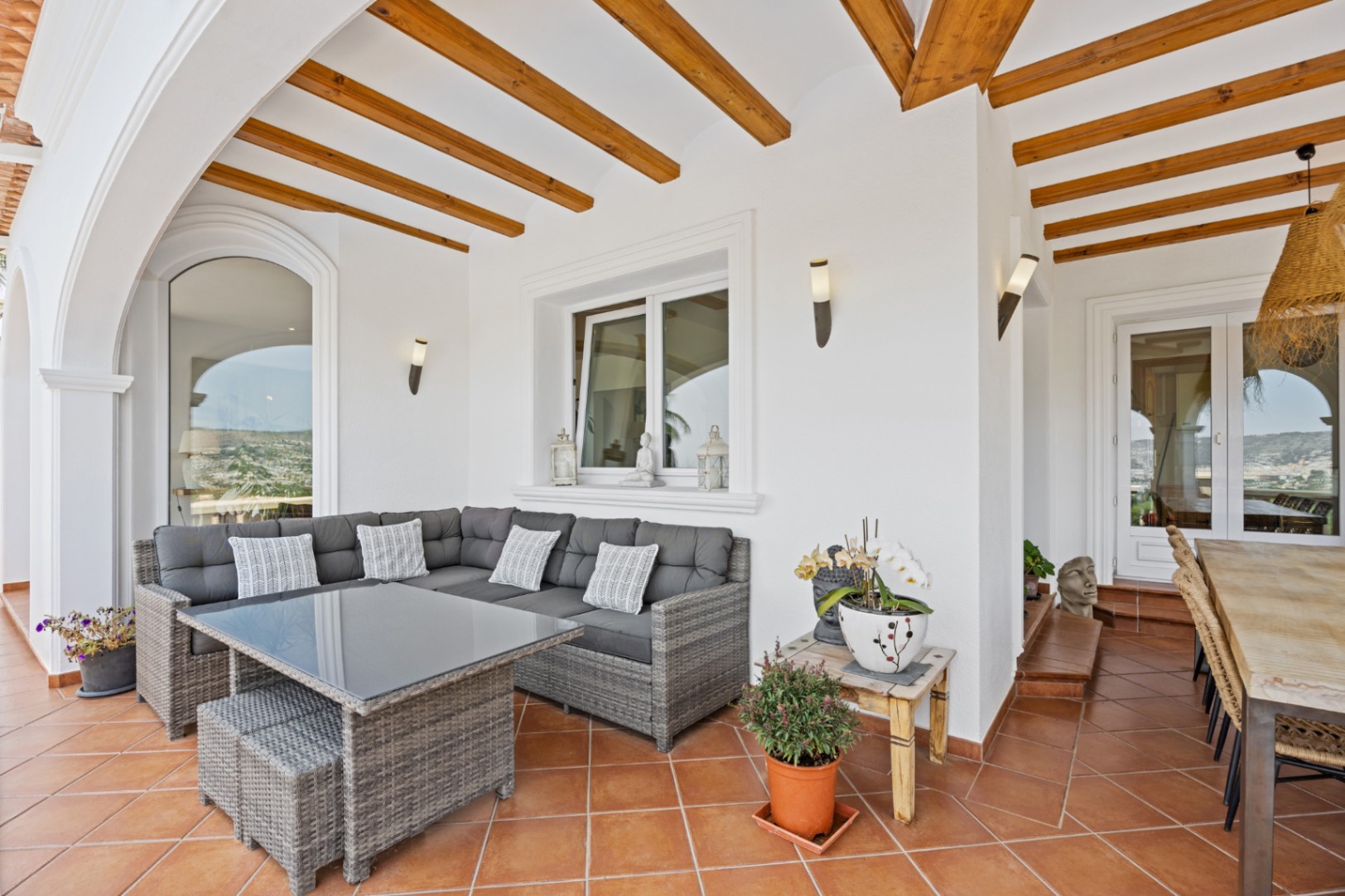 Terrasse couverte dotée de poutres en bois apparentes, d'un sol en terre cuite et d'un salon en résine tressée grise avec table à plateau en verre.