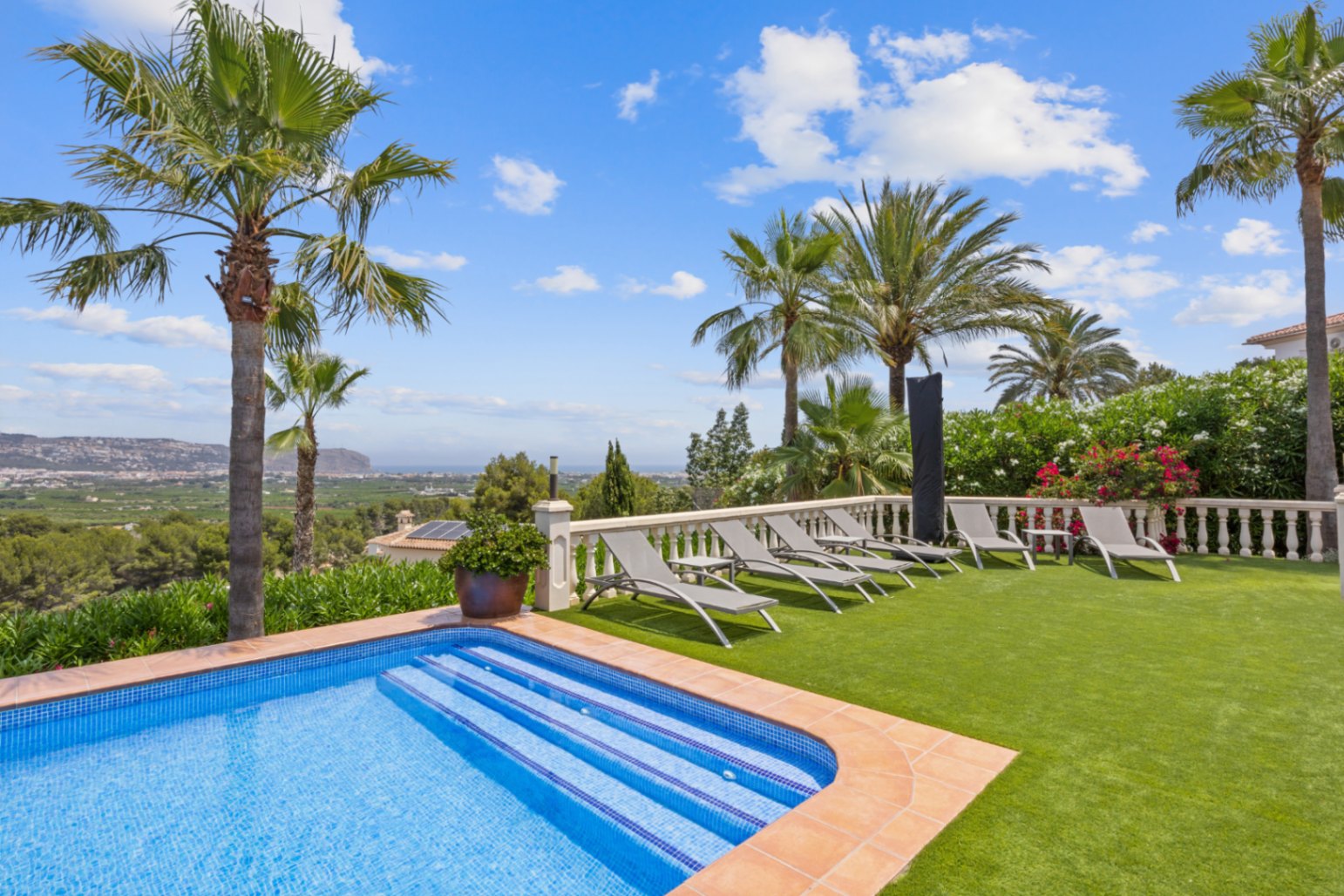 Terrasse de piscine avec chaises longues grises sur gazon synthétique, balustrade en pierre blanche et vue panoramique sur la vallée et la mer.