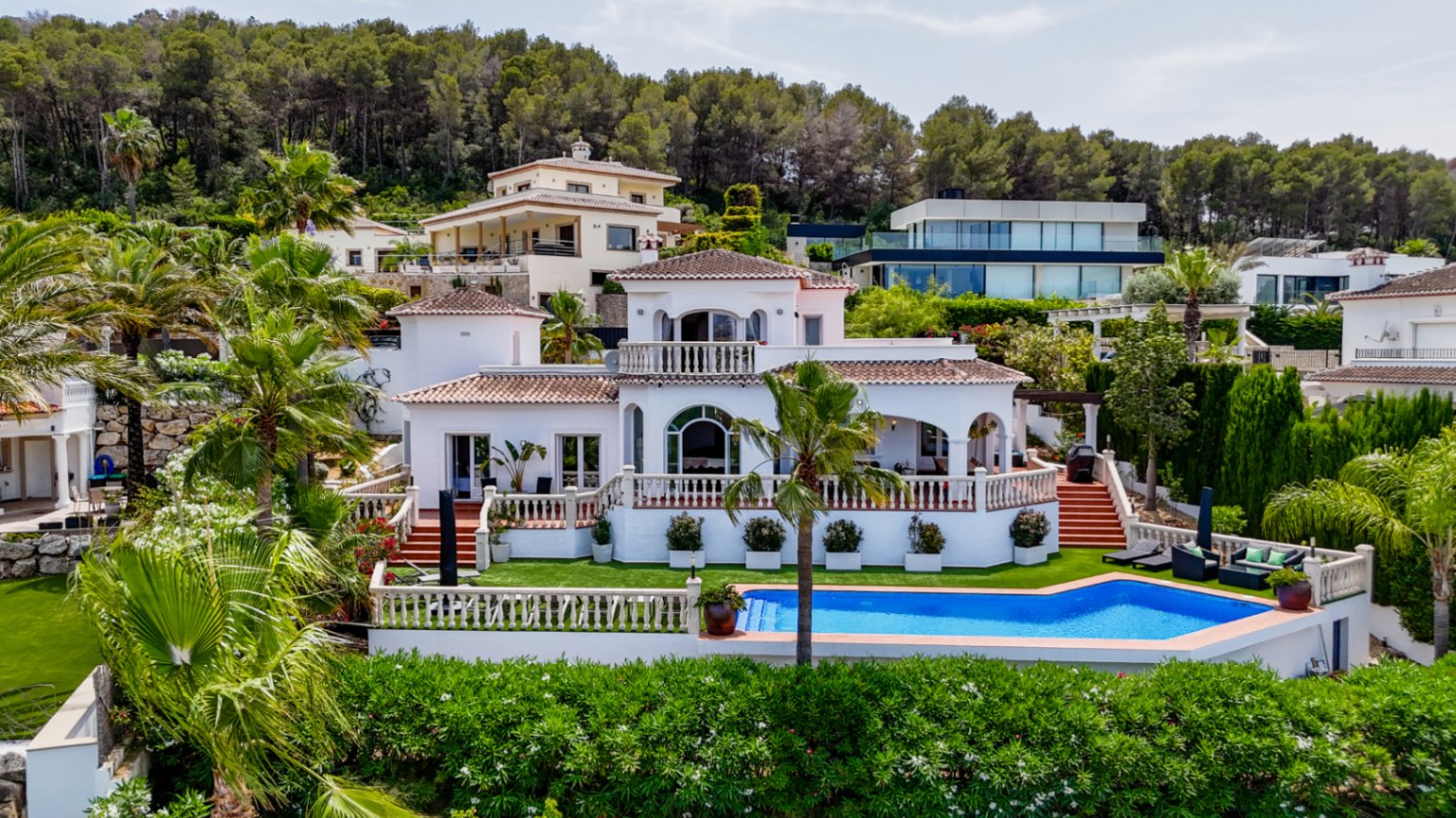Villa méditerranéenne avec façade blanche, toits en terre cuite, terrasses à balustrades et piscine privée, nichée dans un paysage de collines boisées.
