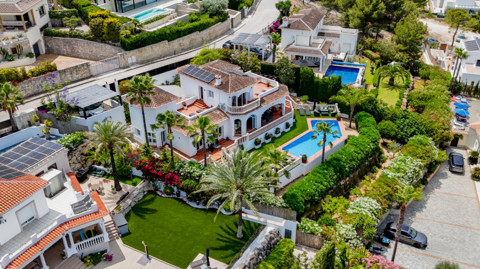 Vue aérienne d'une villa méditerranéenne avec toits en terre cuite, panneaux solaires, piscine privée géométrique et jardins paysagers avec palmiers.