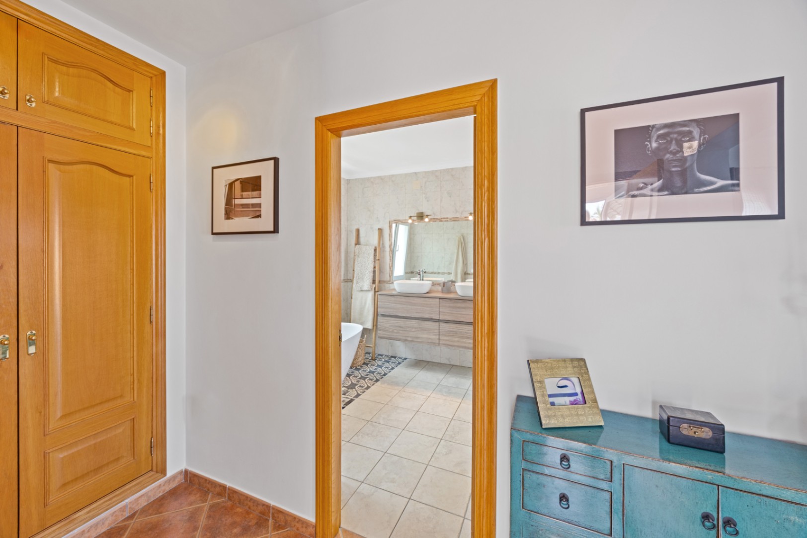 Vue d'un couloir avec placards intégrés en bois et buffet bleu, menant à une salle de bain équipée d'une double vasque et de finitions carrelées.