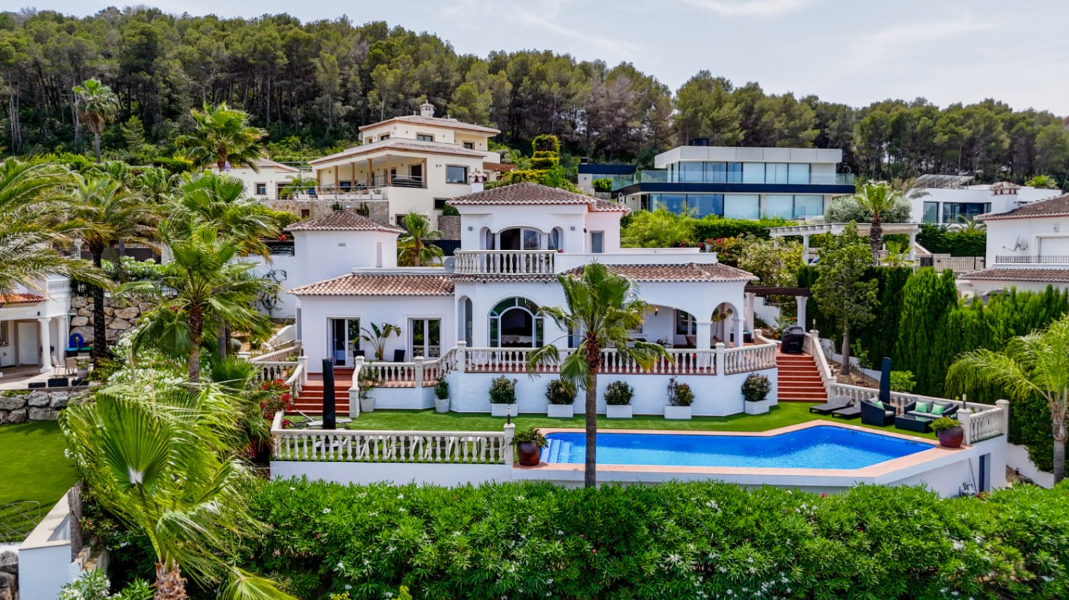 Vue surélevée d'une villa méditerranéenne blanche avec toits en terre cuite, fenêtres cintrées et piscine privée entourée de palmiers et d'une pinède.