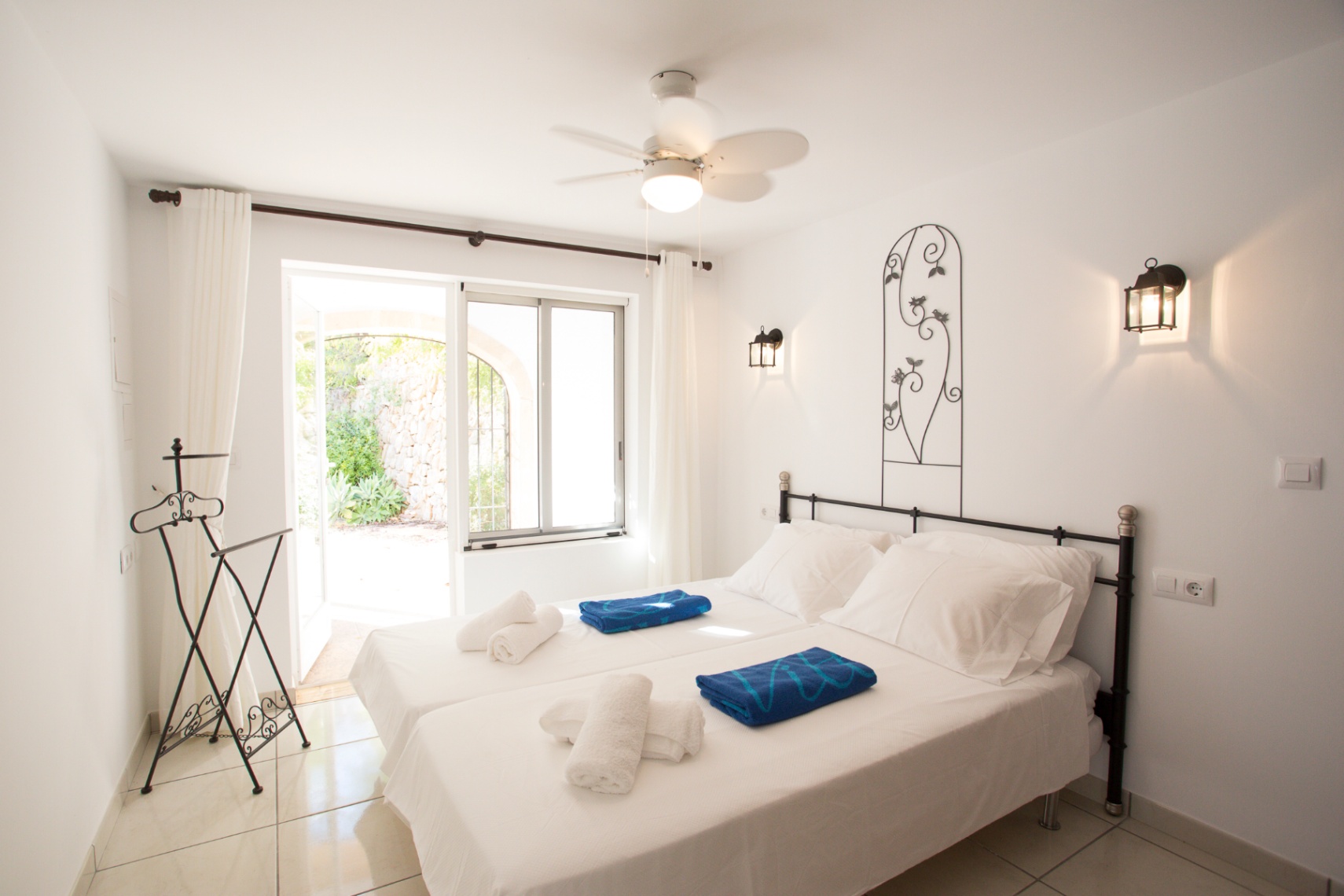 Chambre avec lits jumeaux, tête de lit en fer forgé et appliques murales. Comprend un ventilateur de plafond et un accès direct à un patio extérieur en pierre.