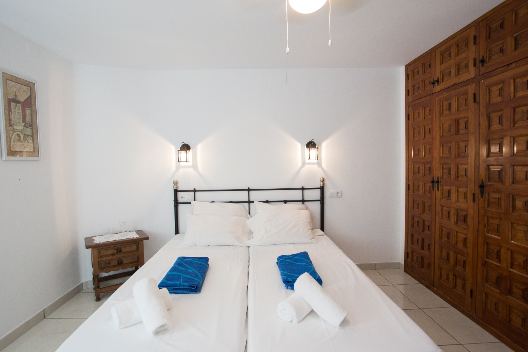 Chambre avec lits jumeaux, tête de lit en fer forgé noir, appliques lanternes et une grande armoire encastrée en bois aux panneaux sculptés.