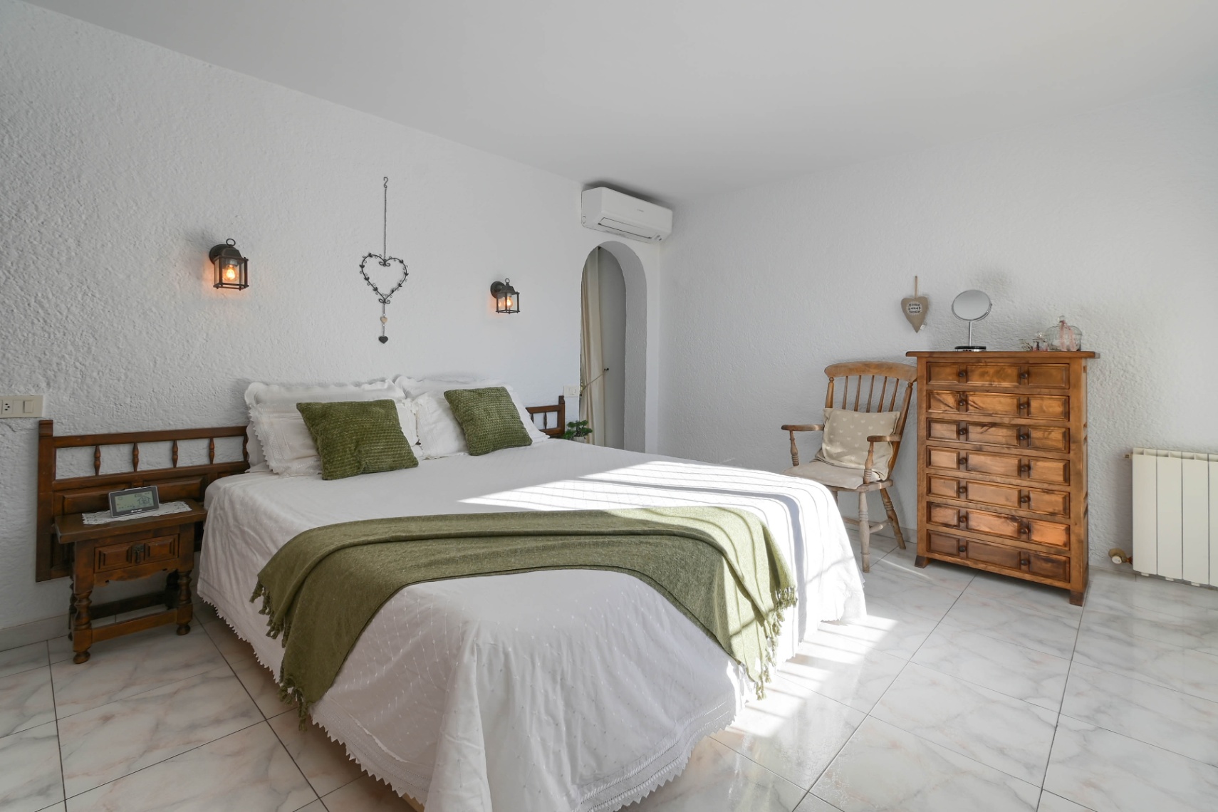 Chambre avec murs texturés blancs, sol en marbre et mobilier en bois rustique. Éclairage par lanternes murales et textiles aux accents verts.