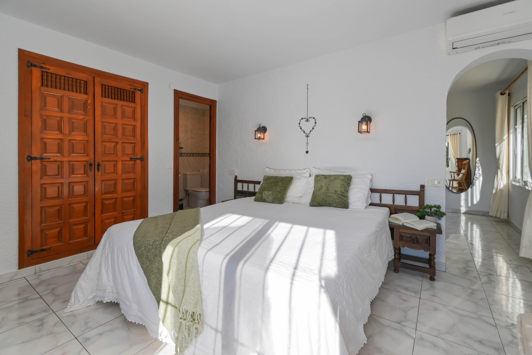 Chambre avec placard intégré en bois sculpté, sol en marbre clair et salle de bain attenante. La lumière naturelle éclaire le lit aux tons verts et les appliques de style lanterne.