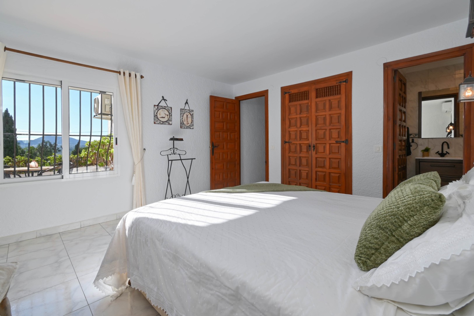 Chambre avec placards intégrés en bois traditionnel, sol en carrelage effet marbre et accès à une salle de bain attenante. La fenêtre offre une vue sur les montagnes.
