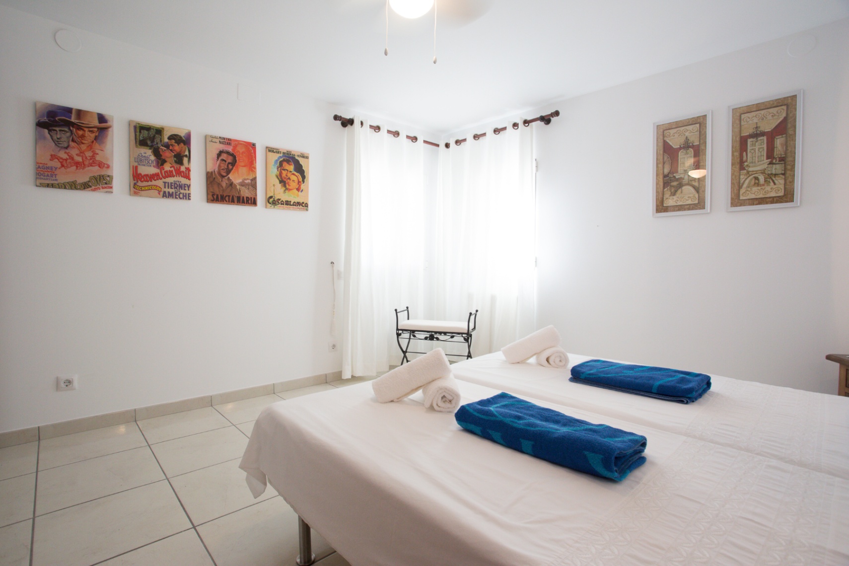 Chambre lumineuse avec lits jumeaux, affiches de cinéma vintage, sol en carrelage et fenêtres d'angle avec rideaux blancs.