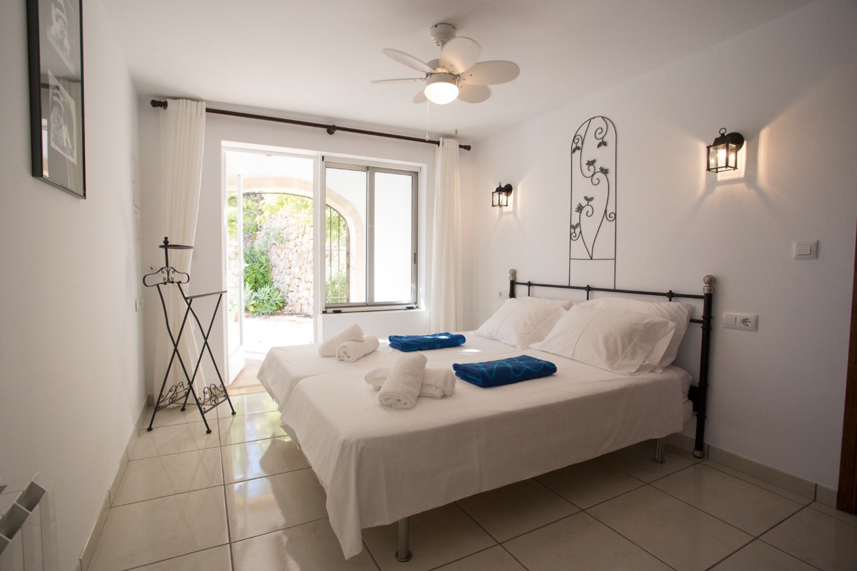 Chambre lumineuse avec lits jumeaux, fer forgé et ventilateur de plafond. Une porte vitrée s'ouvre sur un patio privé aux murs en pierre naturelle.