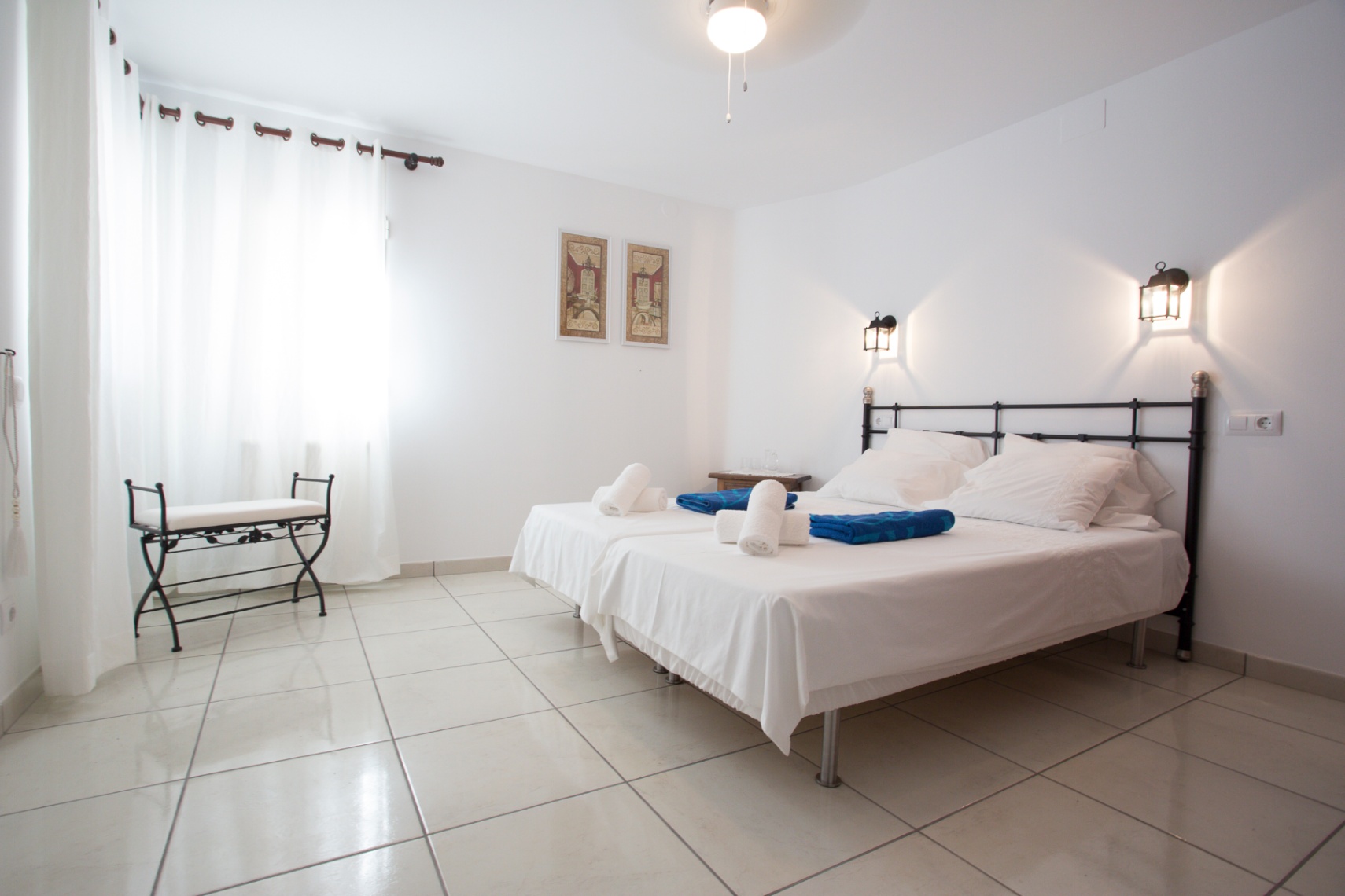 Chambre lumineuse avec lits jumeaux, tête de lit en fer forgé et sol carrelé. Équipée d'appliques murales style lanterne et d'une banquette en fer assortie.