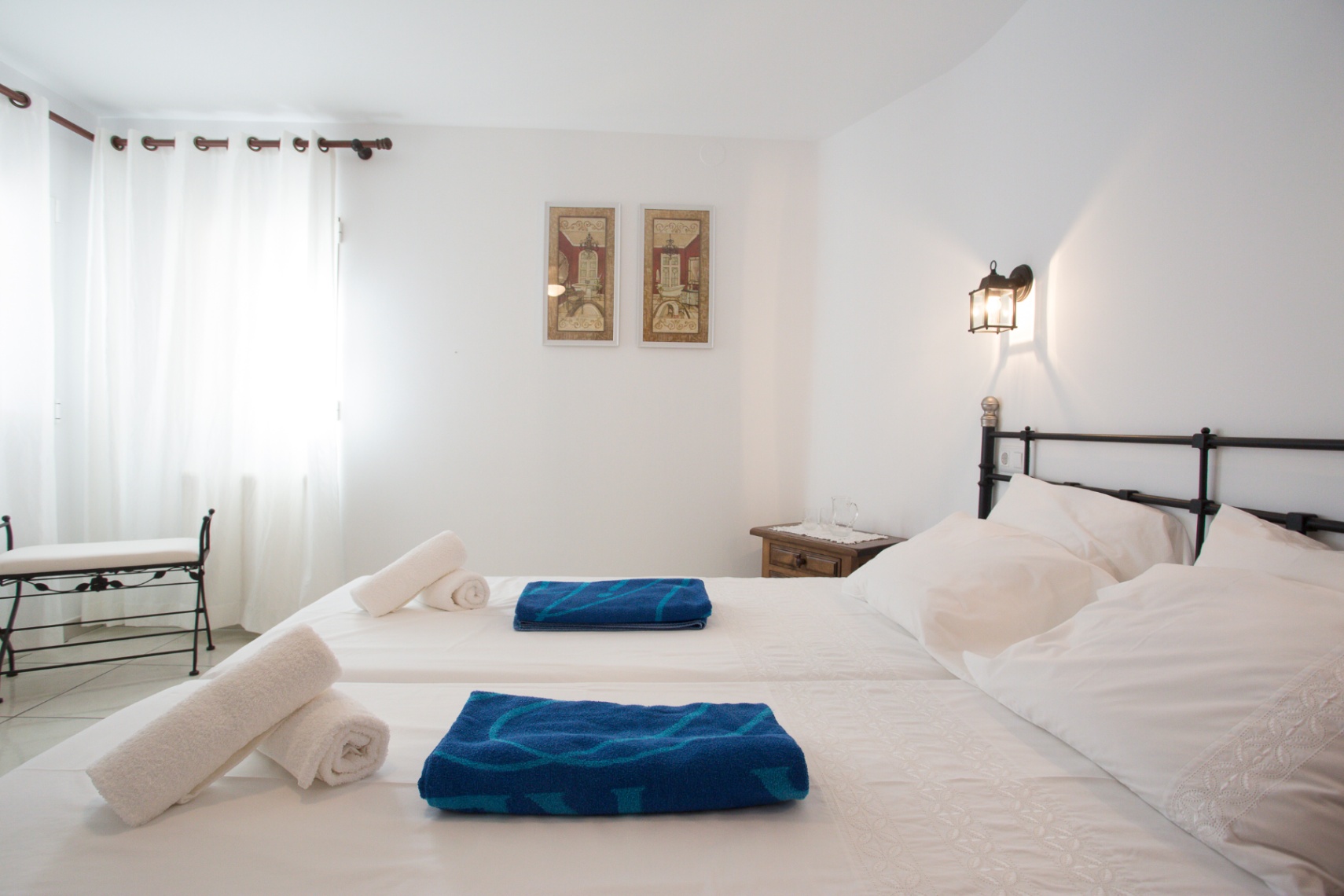 Chambre lumineuse avec lits jumeaux, tête de lit en fer forgé noir et banc assorti. La pièce dispose d'une table de chevet en bois et d'un éclairage naturel.