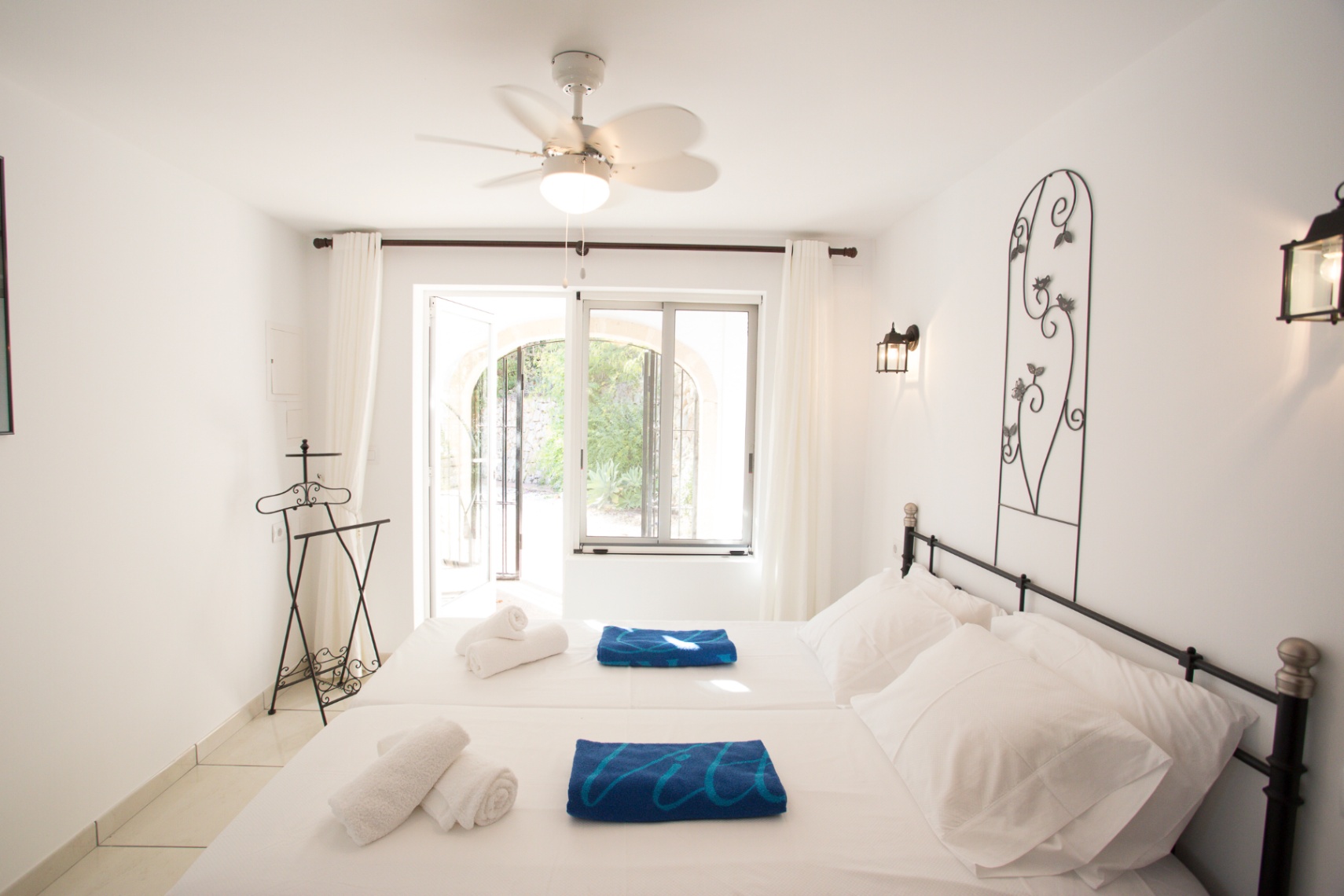 Chambre lumineuse avec murs blancs, tête de lit en fer forgé et ventilateur de plafond. Une porte vitrée s'ouvre sur un patio verdoyant.