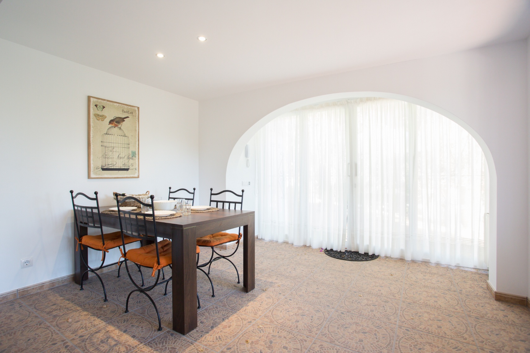 Coin repas avec table en bois sombre et chaises en fer forgé. Une grande ouverture en arche avec voilages blancs illumine le sol en carrelage à motifs.