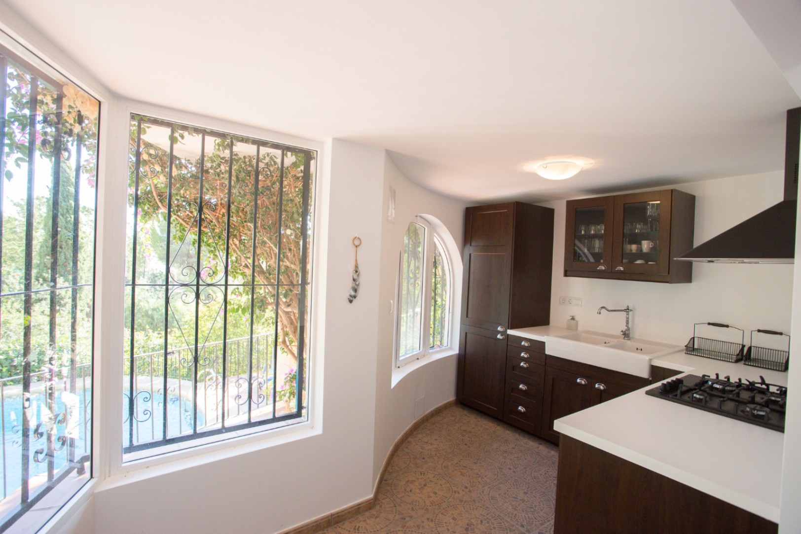 Cuisine dotée de meubles en bois sombre, de plans de travail blancs et d'un évier timbre d'office. Les fenêtres avec ferronnerie donnent sur le jardin et la piscine.