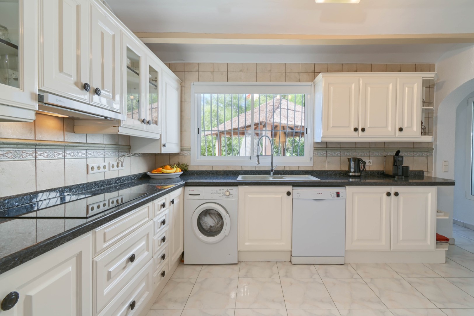 Cuisine fonctionnelle avec meubles blancs, plan de travail en granit foncé et électroménager intégré. Fenêtre lumineuse offrant une vue sur le jardin.