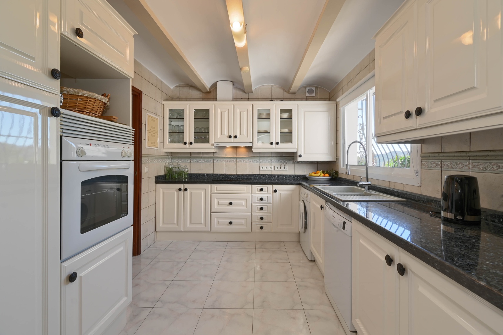 Cuisine traditionnelle équipée de meubles crème, plans de travail en granit sombre et plafond voûté avec poutres apparentes et carrelage.