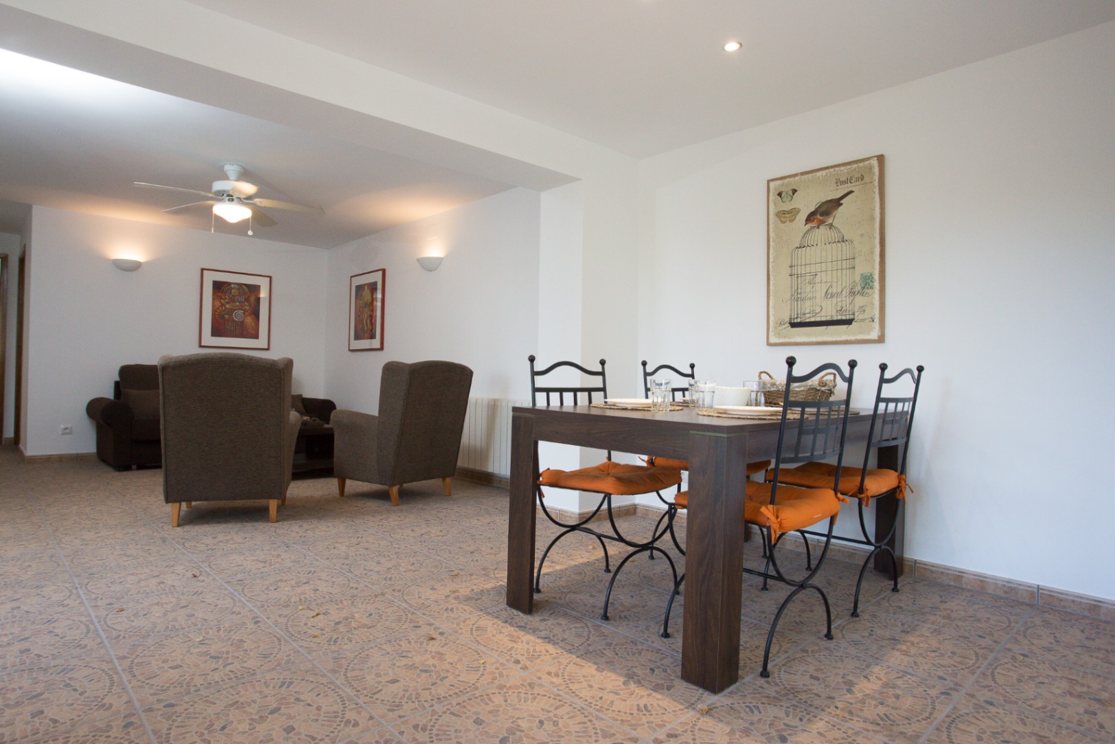 Espace de vie ouvert comprenant une table à manger en bois sombre, des chaises en fer forgé et un coin salon avec fauteuils, le tout sur un sol en carrelage à motifs.