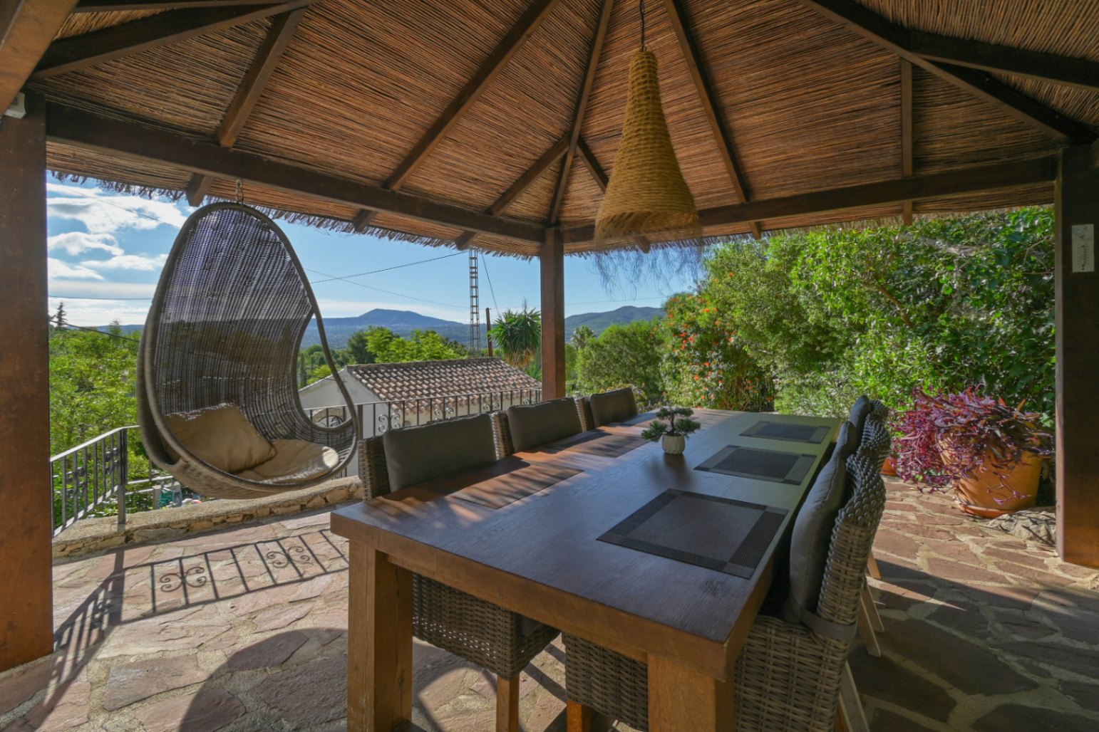 Espace repas extérieur sous un gazebo en chaume avec table en bois, fauteuils en osier et balancelle, offrant une vue dégagée sur les montagnes.