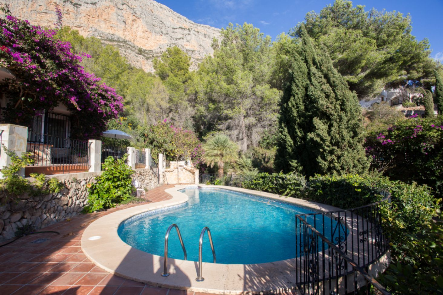 Piscine extérieure de forme libre bordée d'une terrasse en terre cuite et d'une végétation luxuriante, située au pied d'une falaise calcaire.