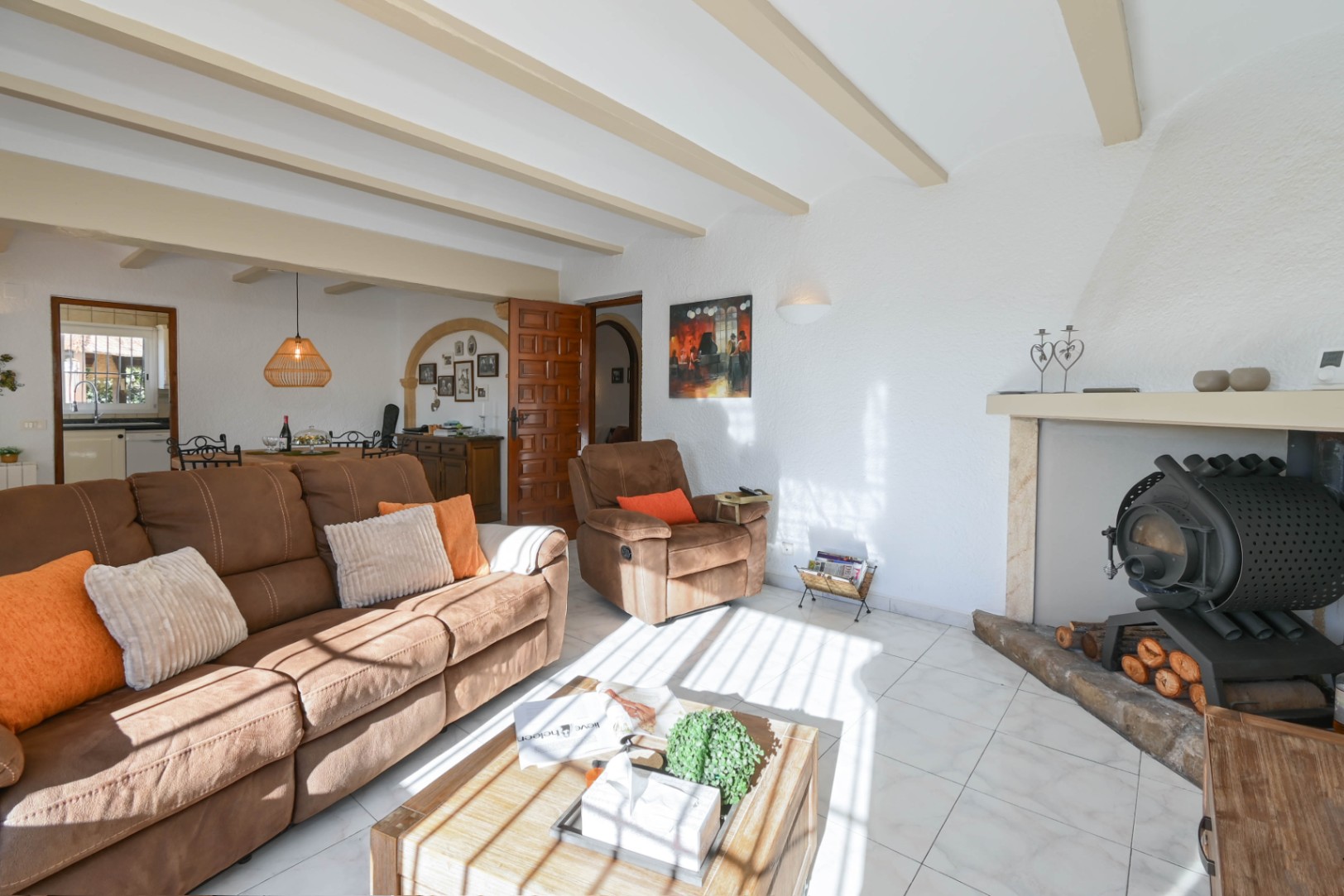 Séjour avec poutres apparentes, canapé marron et poêle à bois traditionnel encastré. La pièce dispose d'un sol en carrelage clair et d'une ouverture sur la salle à manger.