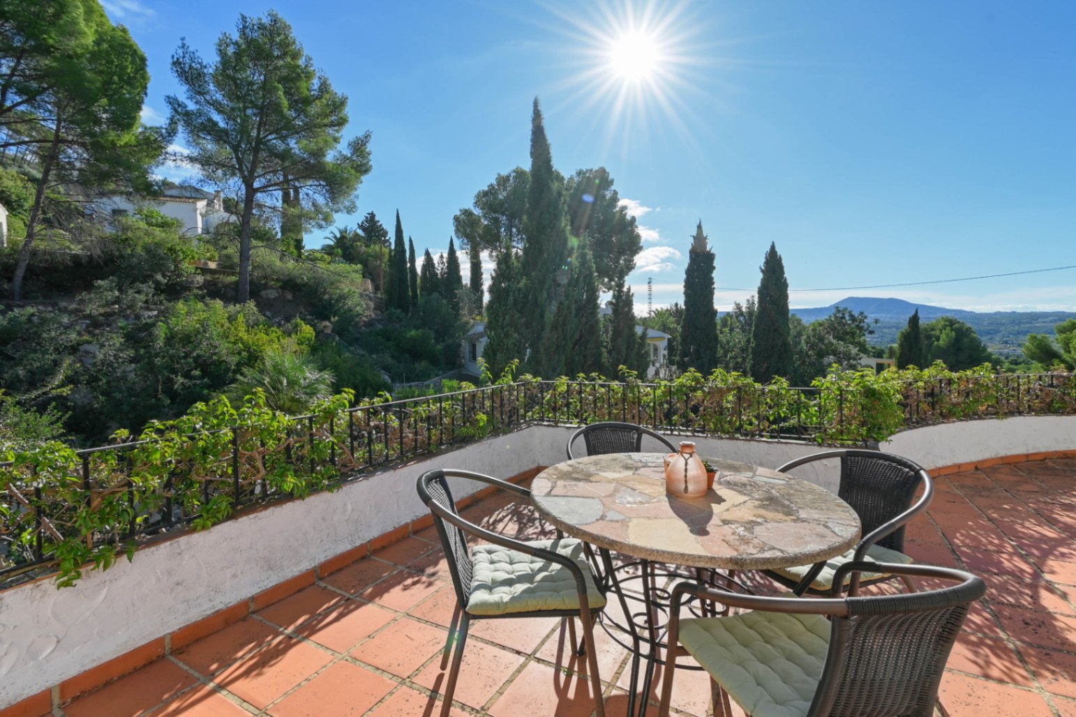 Terrasse avec carrelage en terre cuite, table en pierre et chaises en osier, offrant une vue sur les cyprès et les montagnes sous un soleil éclatant.