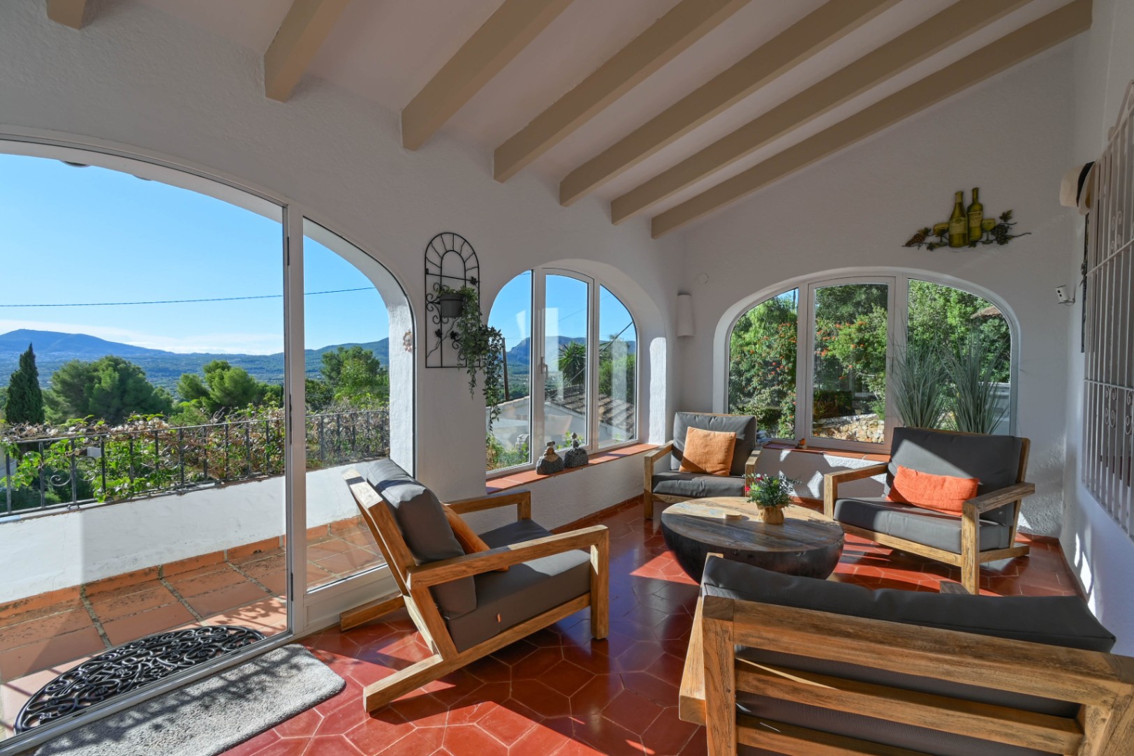 Terrasse couverte avec poutres apparentes, sol en terre cuite hexagonale et baies vitrées cintrées offrant une vue panoramique sur les montagnes.