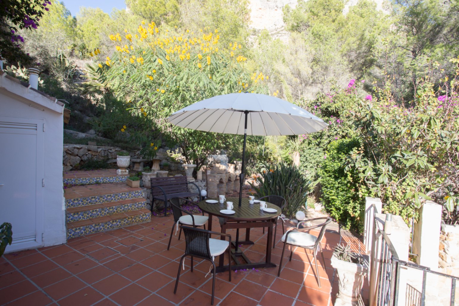 Terrasse en terre cuite avec coin repas sous un parasol blanc, escaliers ornés de carreaux de faïence et vue sur une colline à la végétation méditerranéenne.
