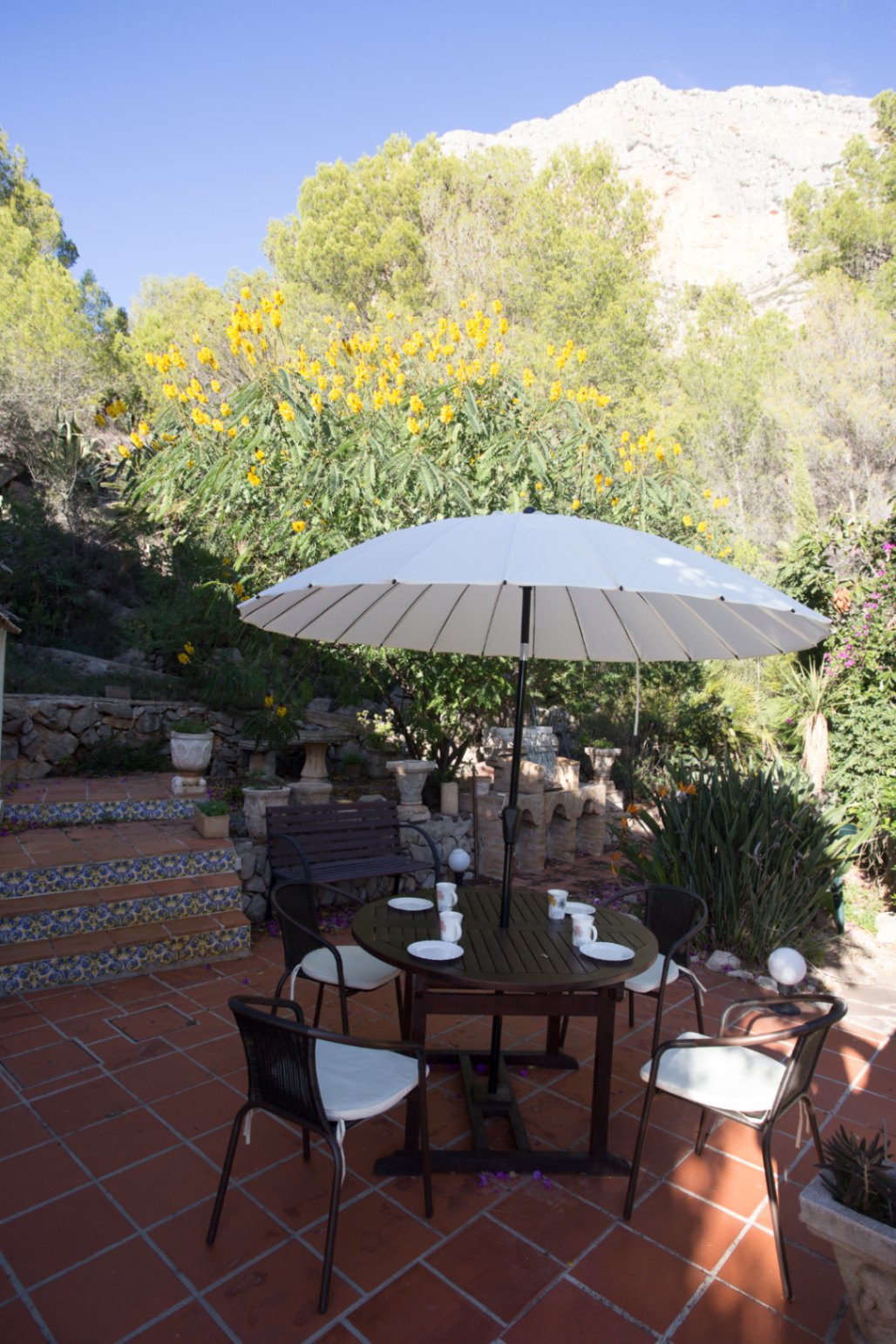 Terrasse extérieure en terre cuite avec table ronde sous parasol blanc et escaliers ornés d'azulejos, offrant une vue sur la montagne et la végétation environnante.