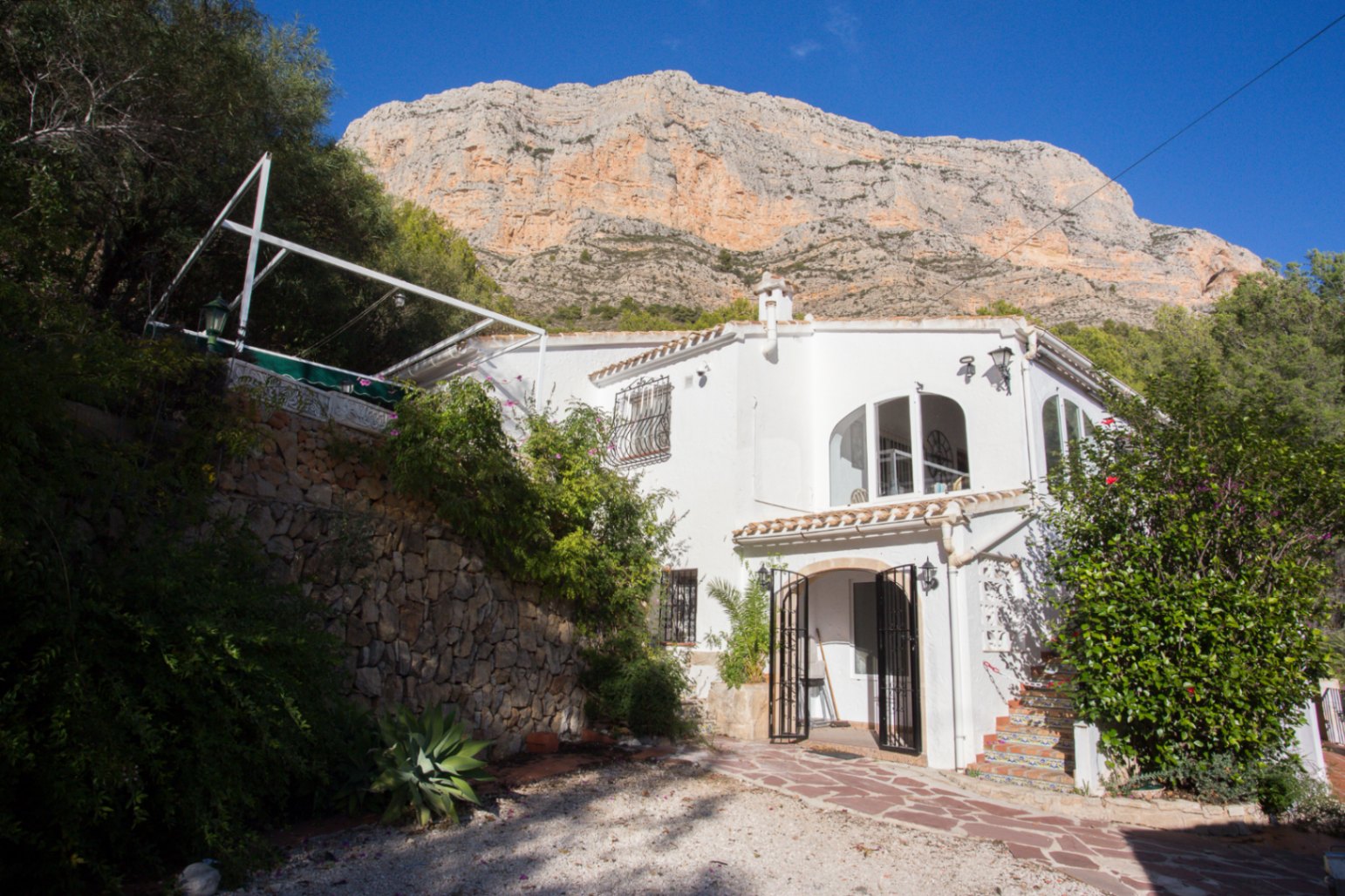Villa méditerranéenne blanche aux fenêtres cintrées, située au pied d'une montagne rocheuse avec des murs en pierre naturelle et un escalier carrelé.