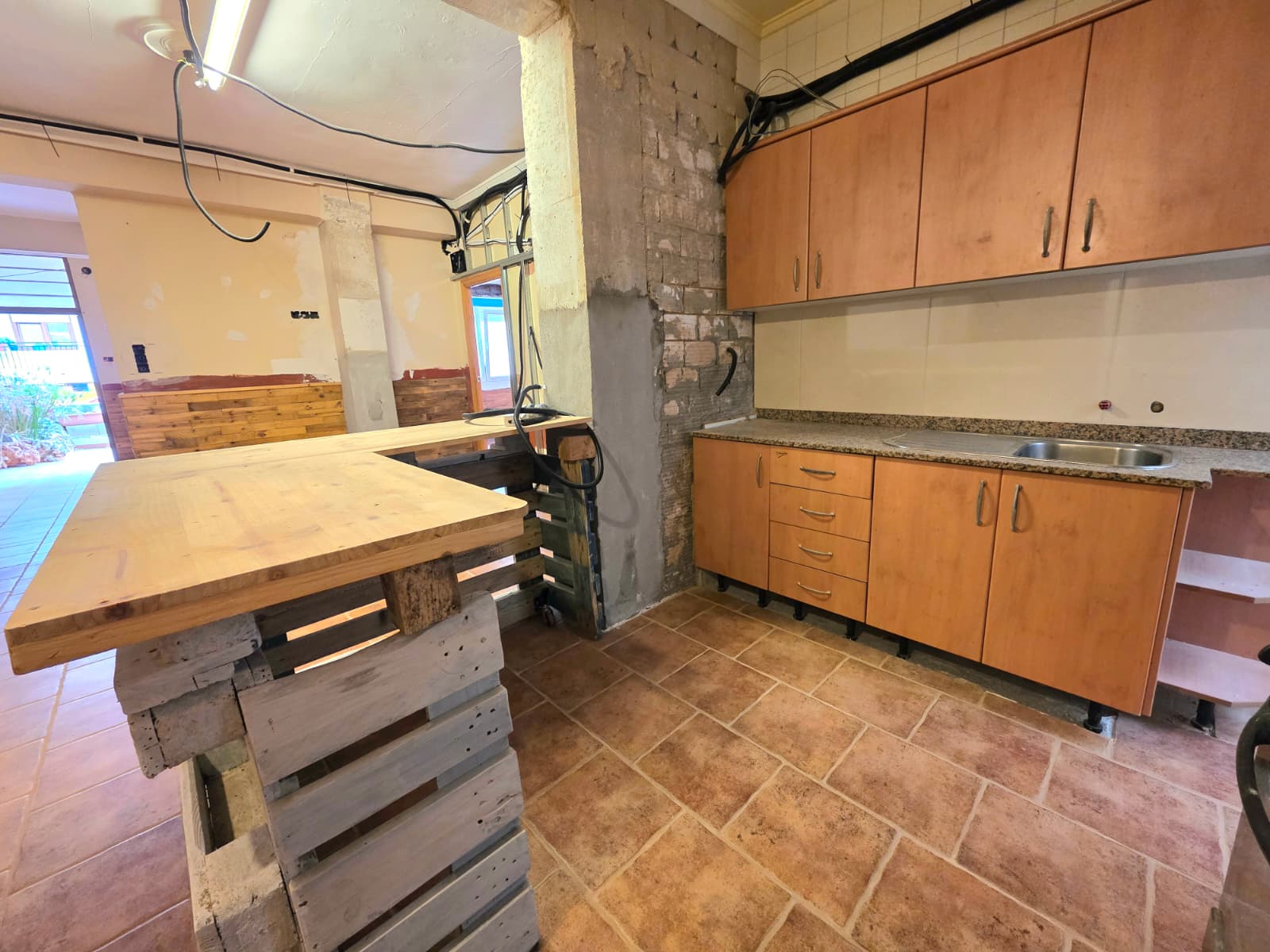 Cuisine en cours de rénovation avec placards en bois, plan de travail en granit, carrelage en terre cuite et un îlot central artisanal en palettes.