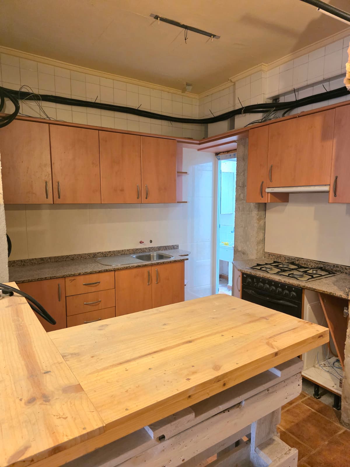 Cuisine équipée de meubles en bois, plans de travail en granit et îlot central en bois brut. Murs carrelés et conduits apparents définissent cet espace.