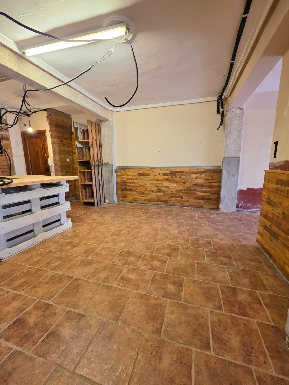 Espace intérieur avec sol en terre cuite, soubassement en bois rustique et un comptoir en palettes blanches surmonté d'un plateau en bois naturel.