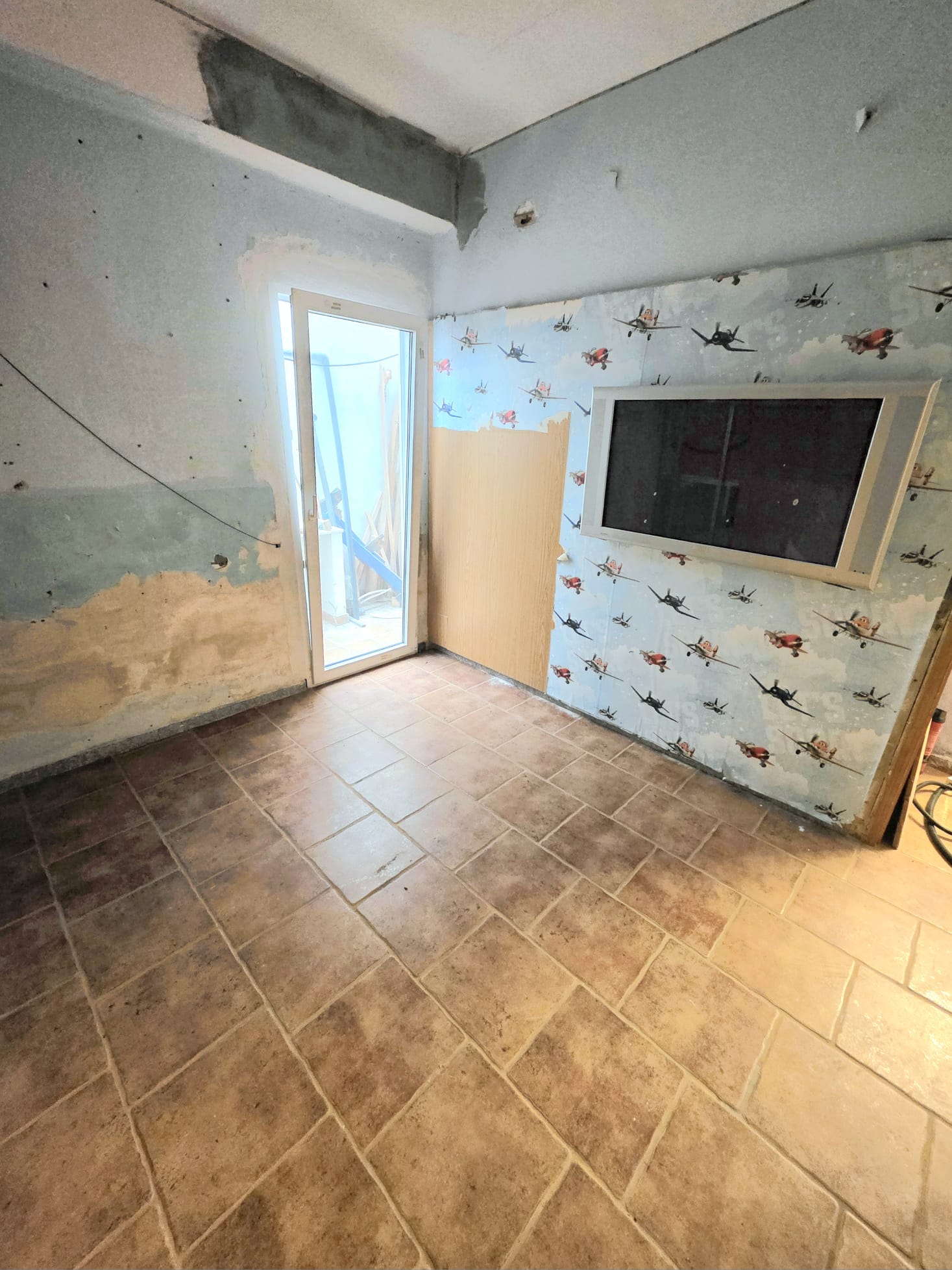 Espace intérieur en cours de rénovation avec sol en grès cérame, porte vitrée à cadre blanc et mur orné d'un papier peint d'avions avec support d'écran intégré.