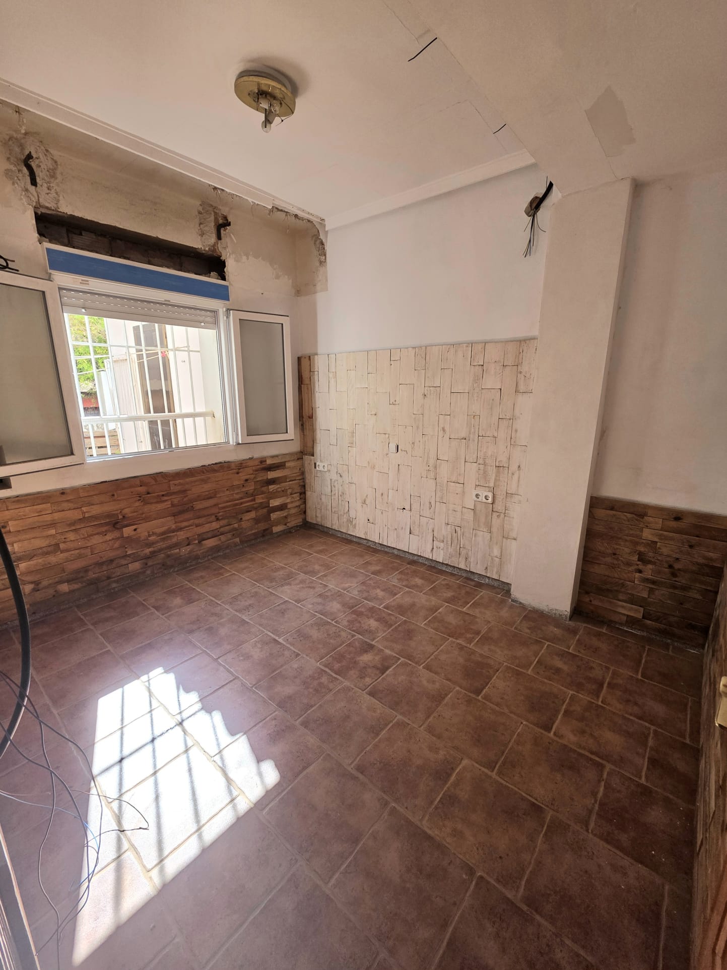 Pièce avec carrelage en terre cuite et revêtement mural en bois décoratif. La lumière naturelle pénètre par la fenêtre, révélant un espace en cours de rénovation.