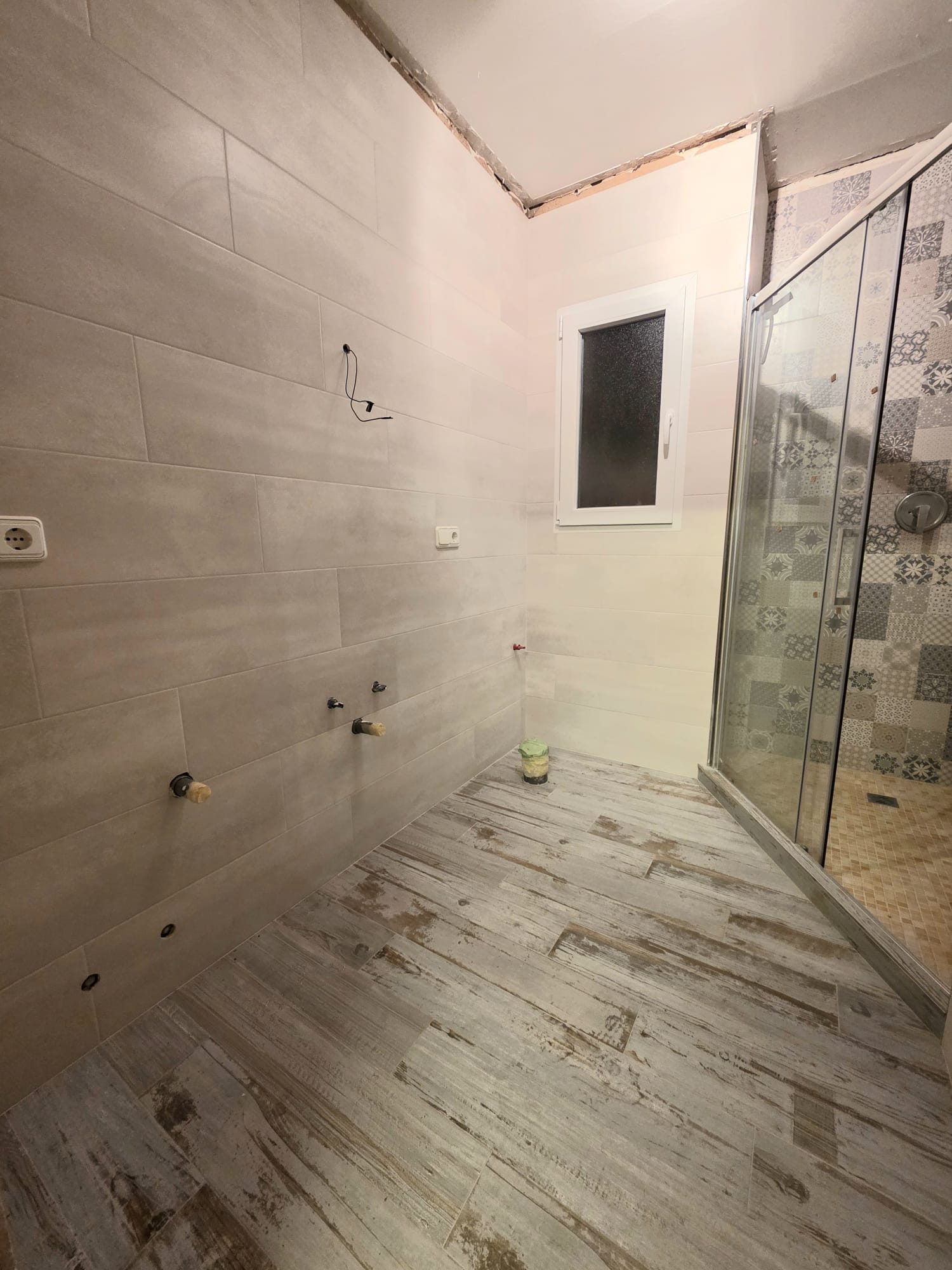 Salle de bain en rénovation avec carrelage imitation bois au sol, murs en grès cérame et douche vitrée ornée de carreaux de ciment à motifs géométriques.