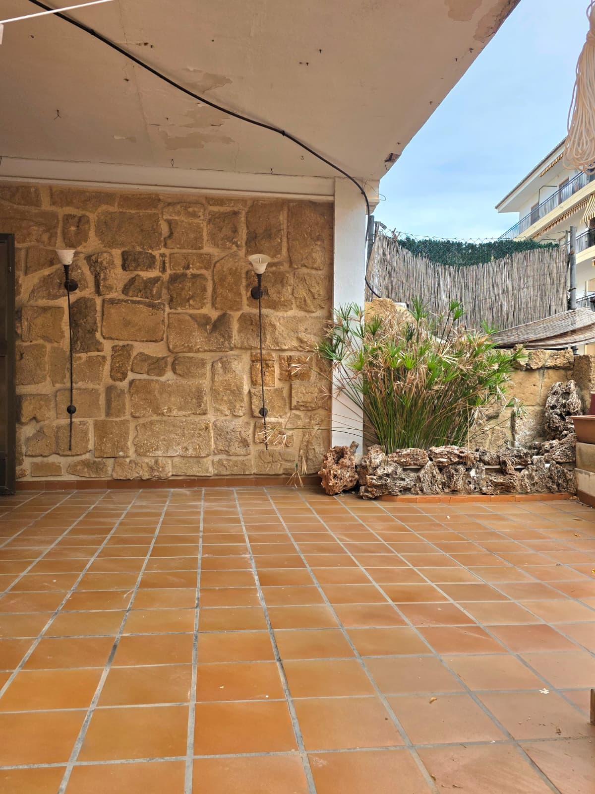 Terrasse couverte avec sol en terre cuite, mur en pierre apparente orné d'appliques murales et jardinière bordée de roches volcaniques.