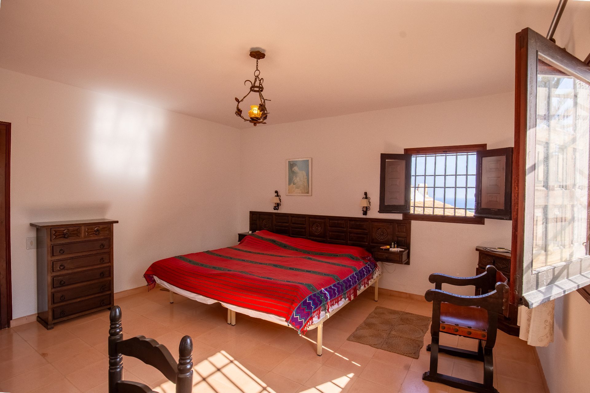 Chambre rustique avec tête de lit en bois sculpté, sol en terre cuite et lustre en fer forgé. Une fenêtre à barreaux et volets en bois apporte une lumière naturelle douce.