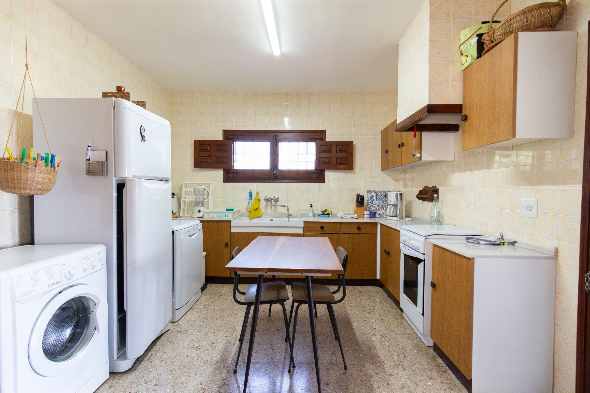 Cuisine fonctionnelle avec meubles en bois, carrelage mural à motifs et électroménagers blancs. Une table à manger centrale repose sur un sol en terrazzo sous une fenêtre à volets.