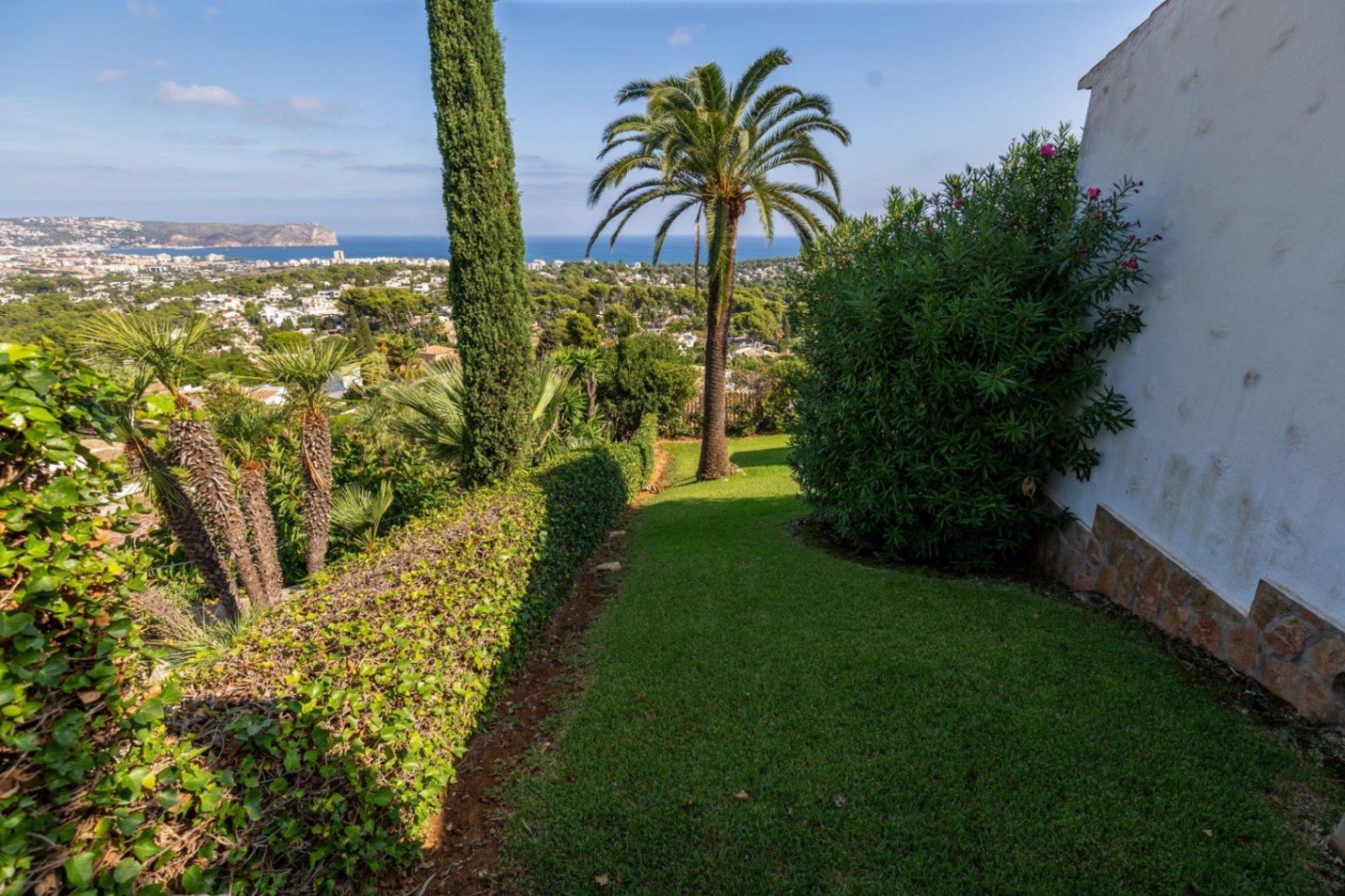 Jardin paysager avec pelouse, palmiers et cyprès, offrant une vue panoramique sur la ville côtière et la mer Méditerranée.
