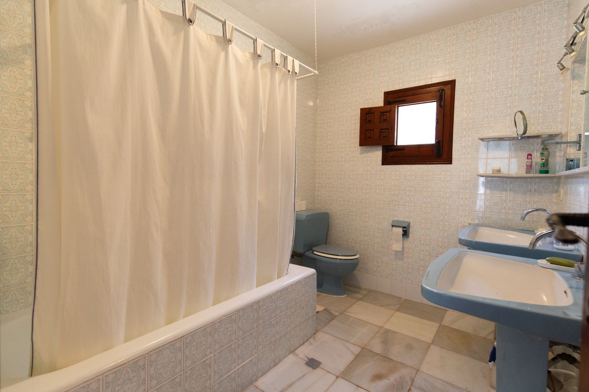 Salle de bain traditionnelle avec double lavabo sur colonne bleu, WC assorti et baignoire carrelée. Murs ornés de carreaux à motifs et petite fenêtre en bois authentique.