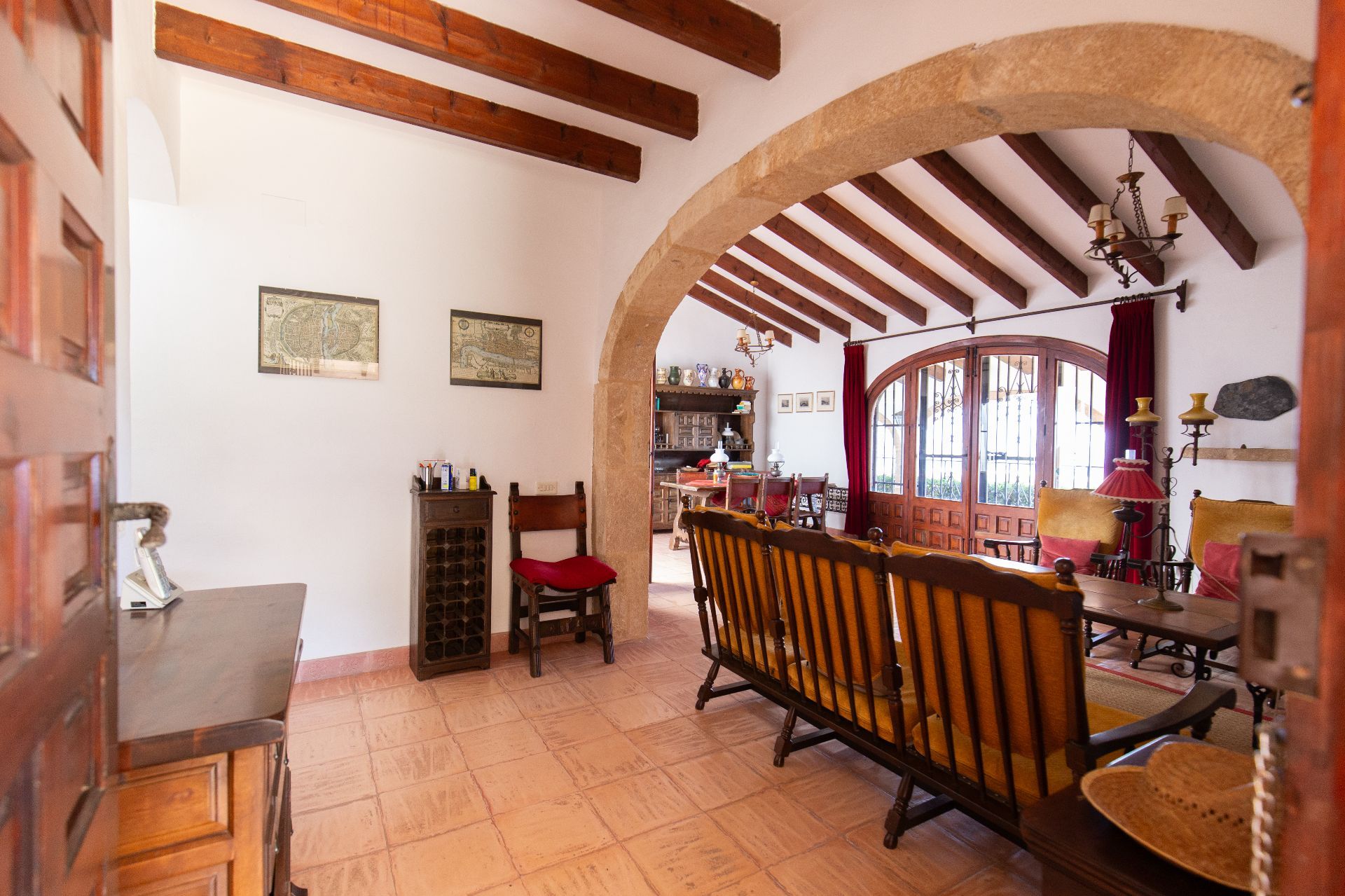 Salon rustique avec poutres en bois apparentes, une arche en pierre et un sol en terre cuite. Des portes cintrées en bois s'ouvrent sur l'extérieur, entourées de meubles classiques.