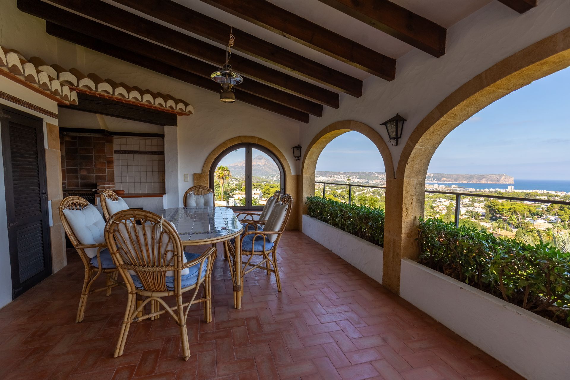 Terrasse couverte avec arches en pierre, poutres apparentes et sol en terre cuite. Espace repas avec vue panoramique sur la mer et la montagne.