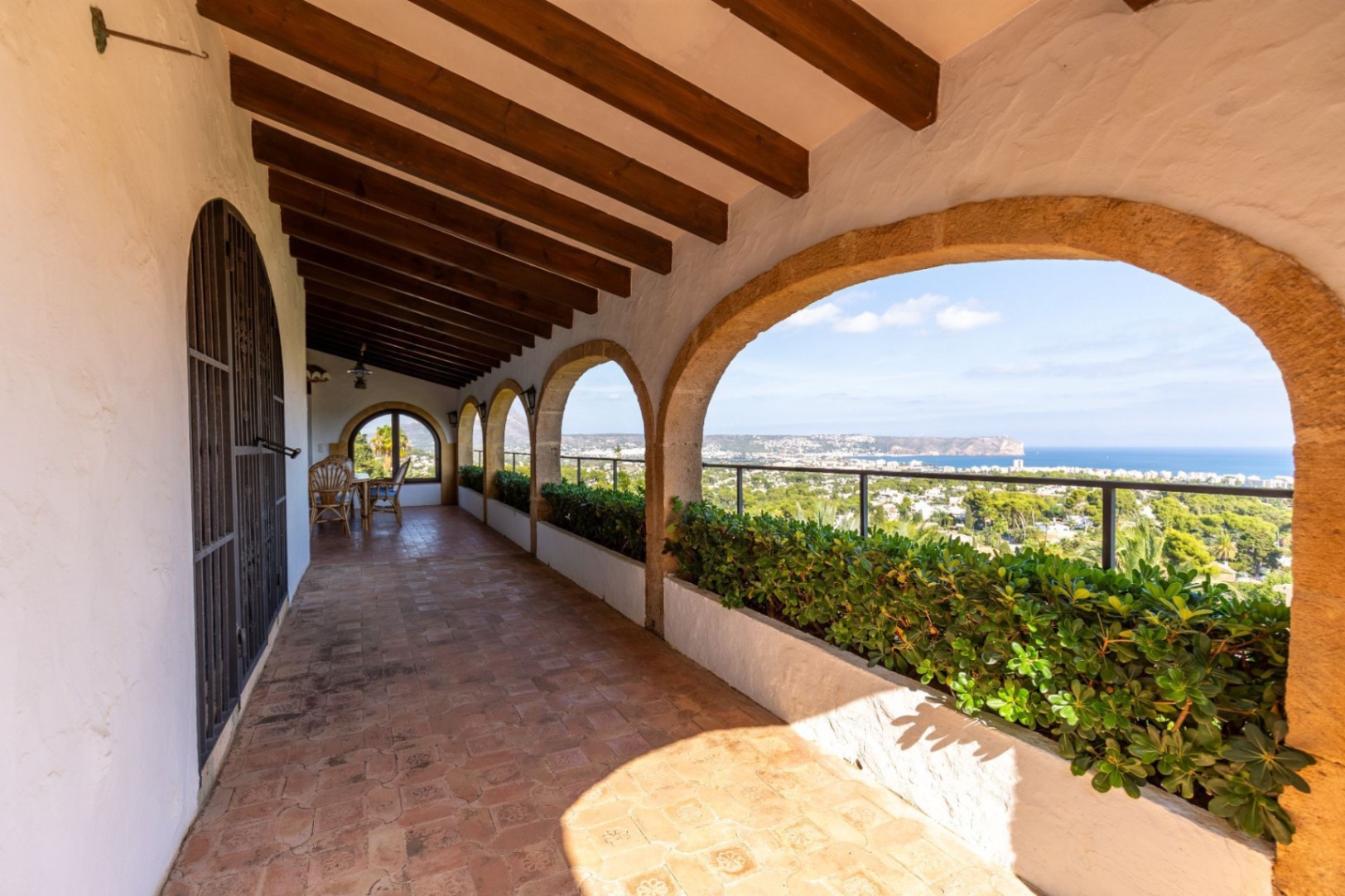 Terrasse couverte avec arches en pierre, poutres en bois apparentes et sol en terre cuite. Des jardinières intégrées offrent une vue panoramique sur la côte et la mer.