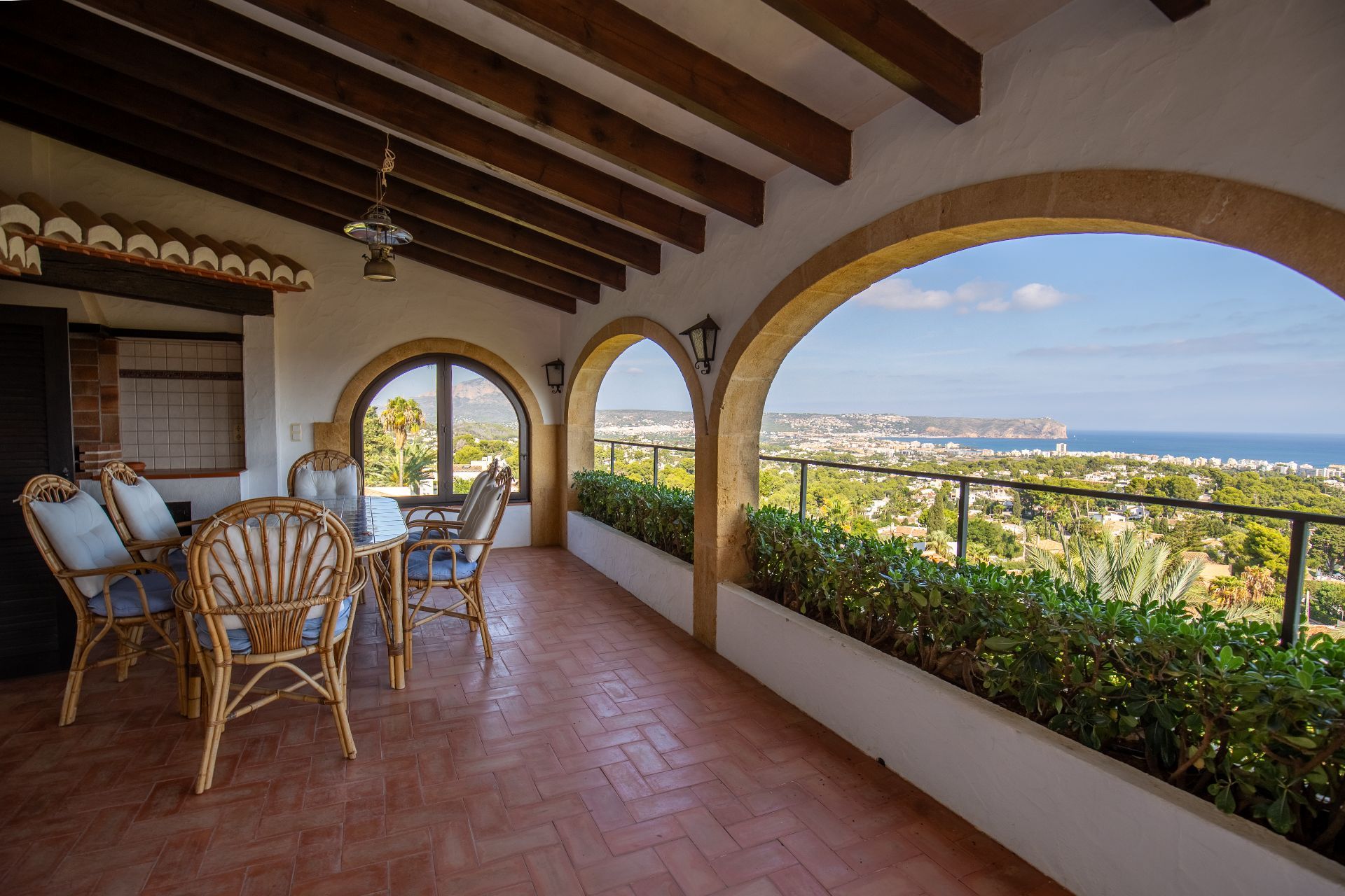 Terrasse couverte avec poutres en bois, sol en terre cuite et arches en pierre offrant une vue panoramique sur la côte et la mer.