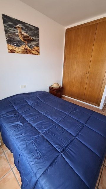 Chambre avec placard intégré en bois, lit avec couette bleue et sol en carrelage terre cuite. Une table de chevet en bois et un tableau marin décorent la pièce.