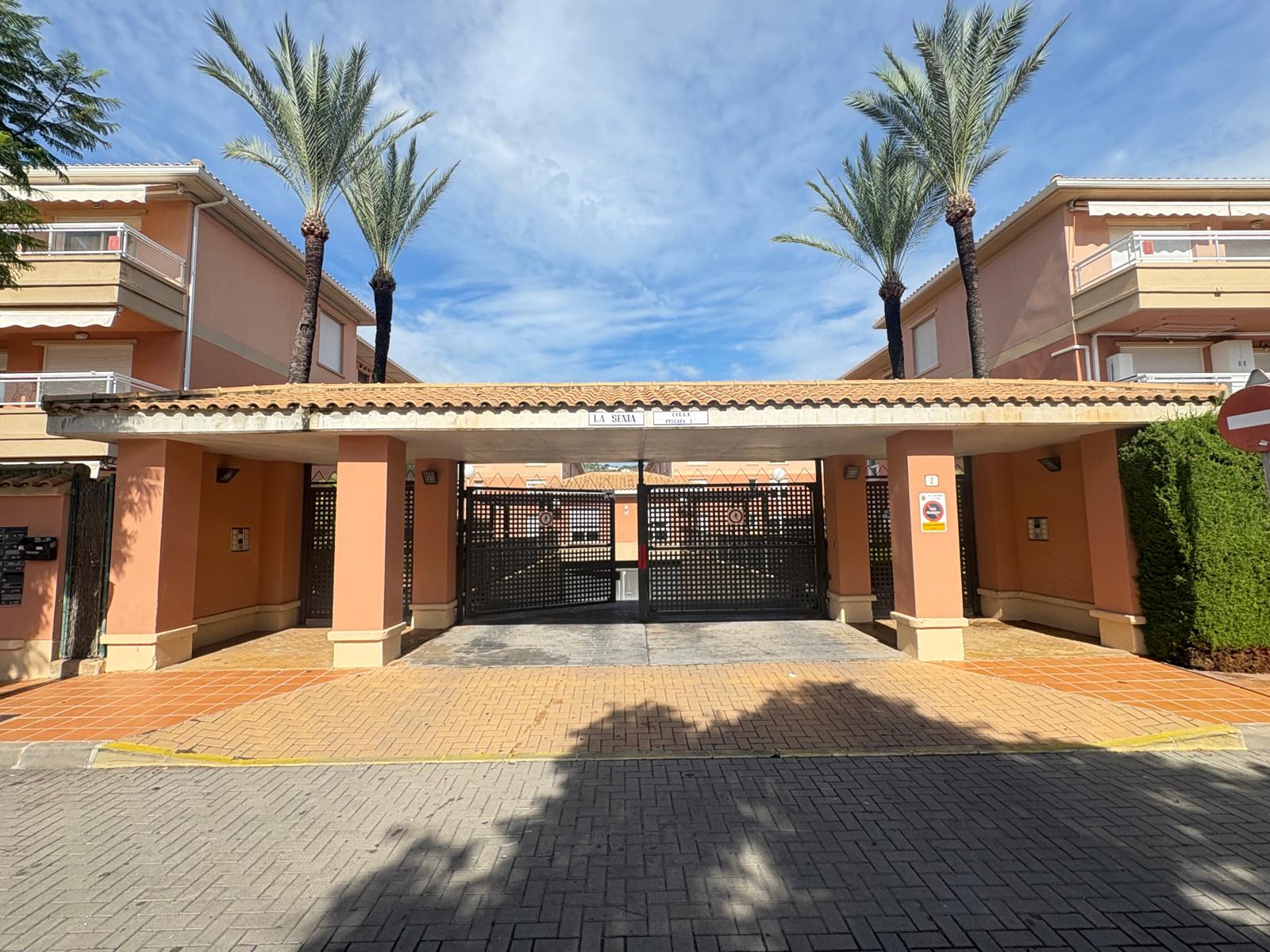 Entrée du complexe résidentiel La Senia avec piliers en terre cuite, toit de tuiles et portails en treillis métallique noir encadrés par des palmiers.