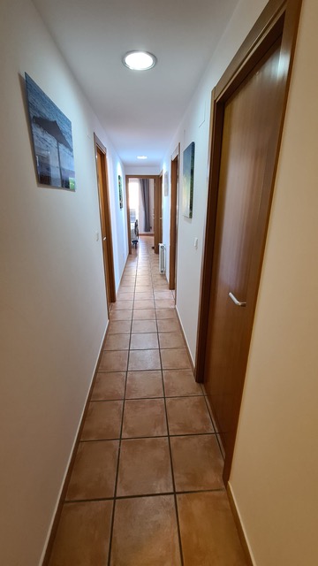 Long couloir avec un sol en carreaux de terre cuite, des portes en bois massif et des murs blancs ornés de photographies et de spots encastrés.