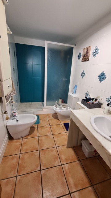 Salle de bain avec douche carrelée en bleu sarcelle, sol en terre cuite et carrelage mural décoratif. Comprend un bidet et un meuble vasque blanc.